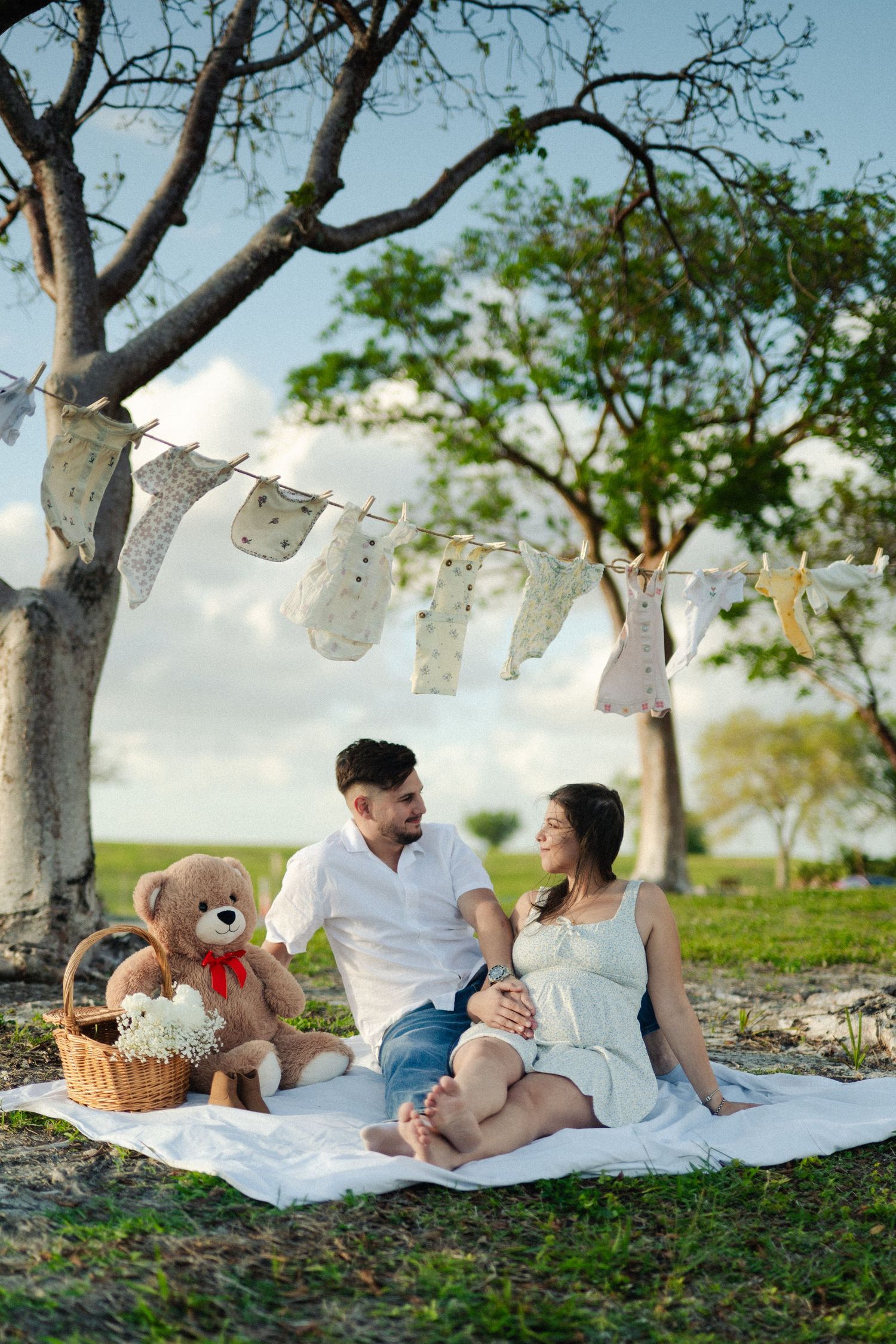Expecting couple sits on a white blanket under a tree with a teddy bear and baby clothes bunting at a park picnic.
