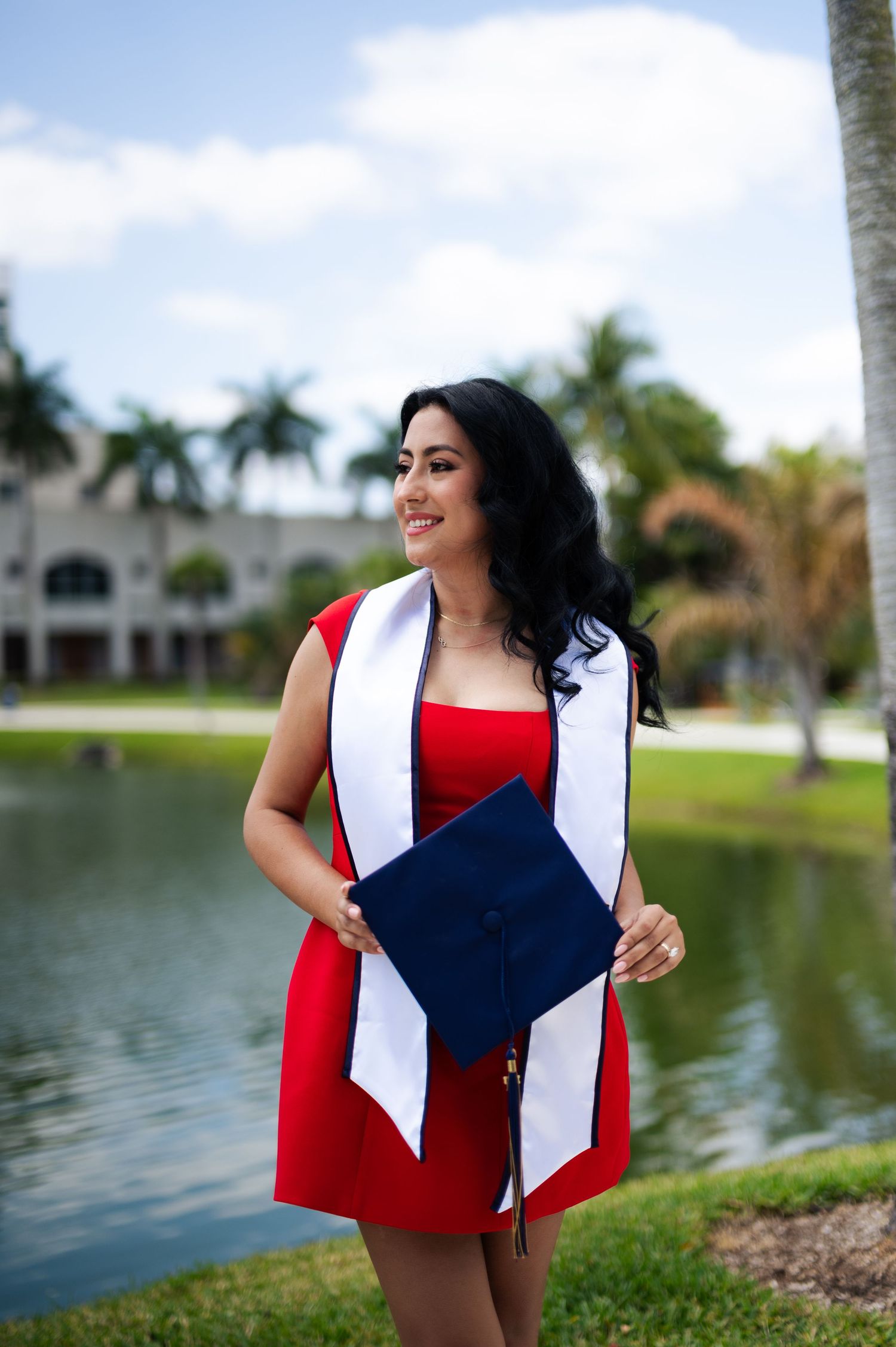 Graduate in a red dress and white sash holds a navy graduation cap beside a lake with palm trees.