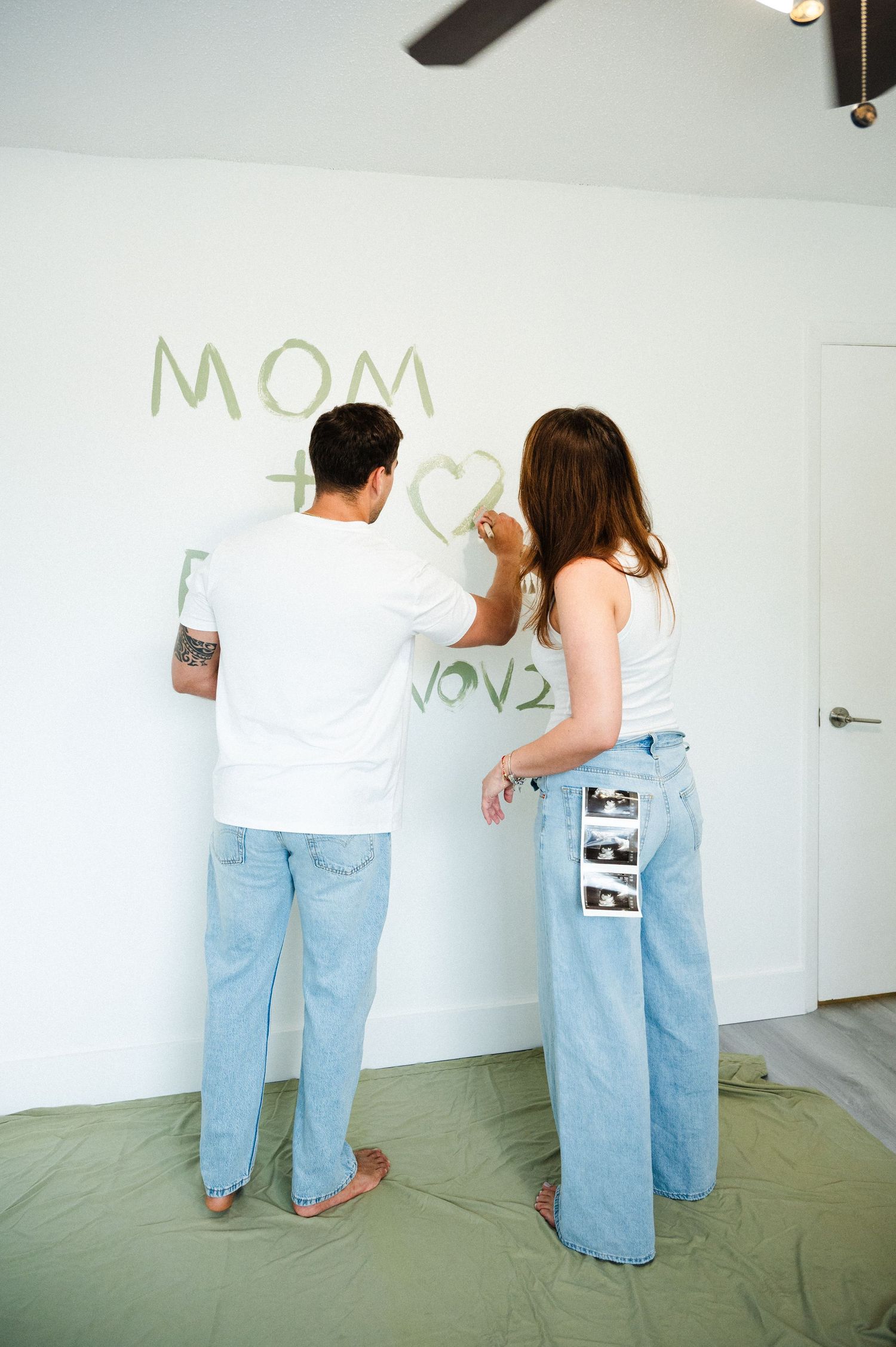 Couple in jeans write a pregnancy announcement in green paint on a white wall, with ultrasound photos in a back pocket.