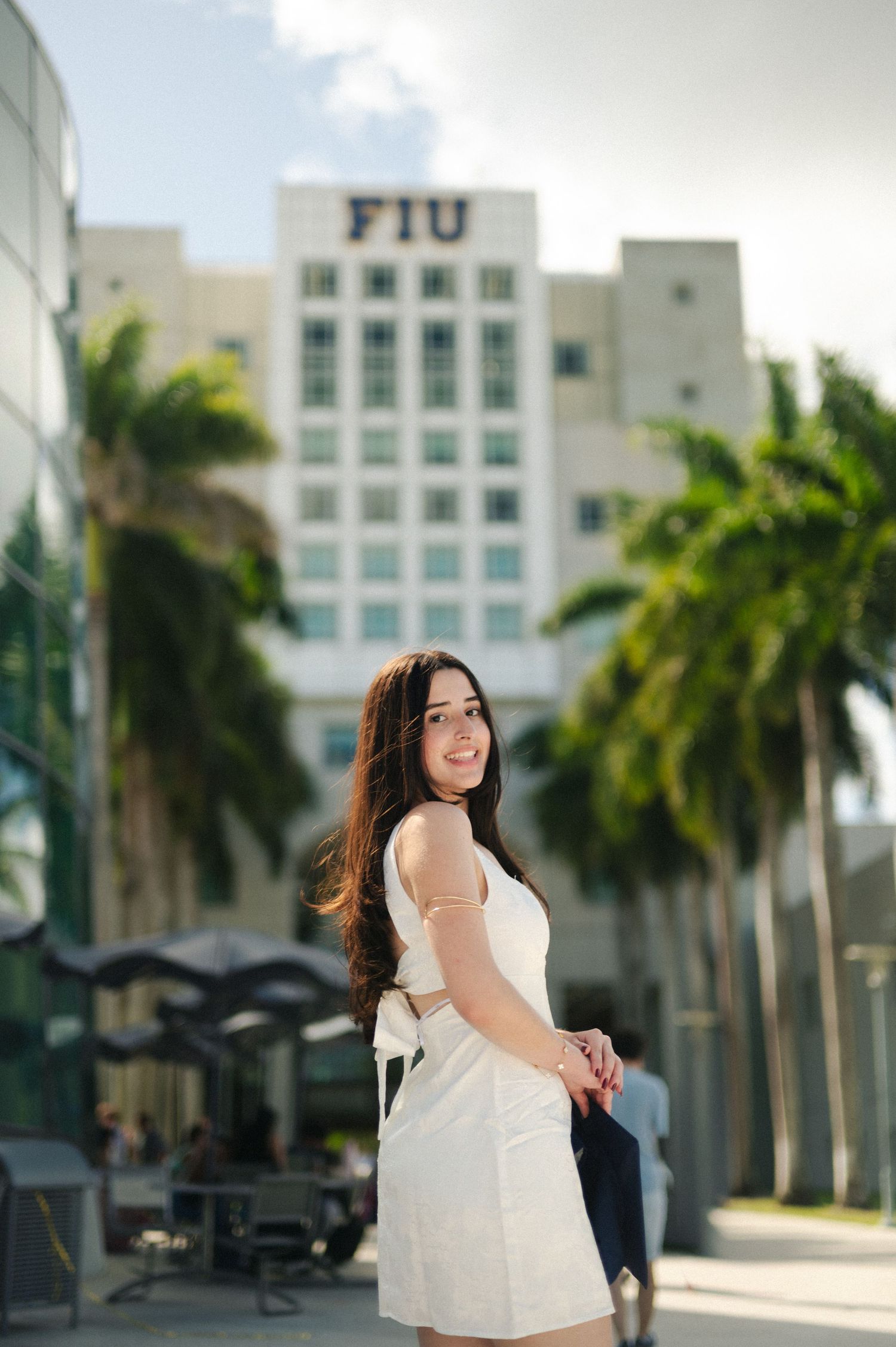 Smiling woman in a white dress poses on a sunny palm-lined walkway in front of an FIU campus building.