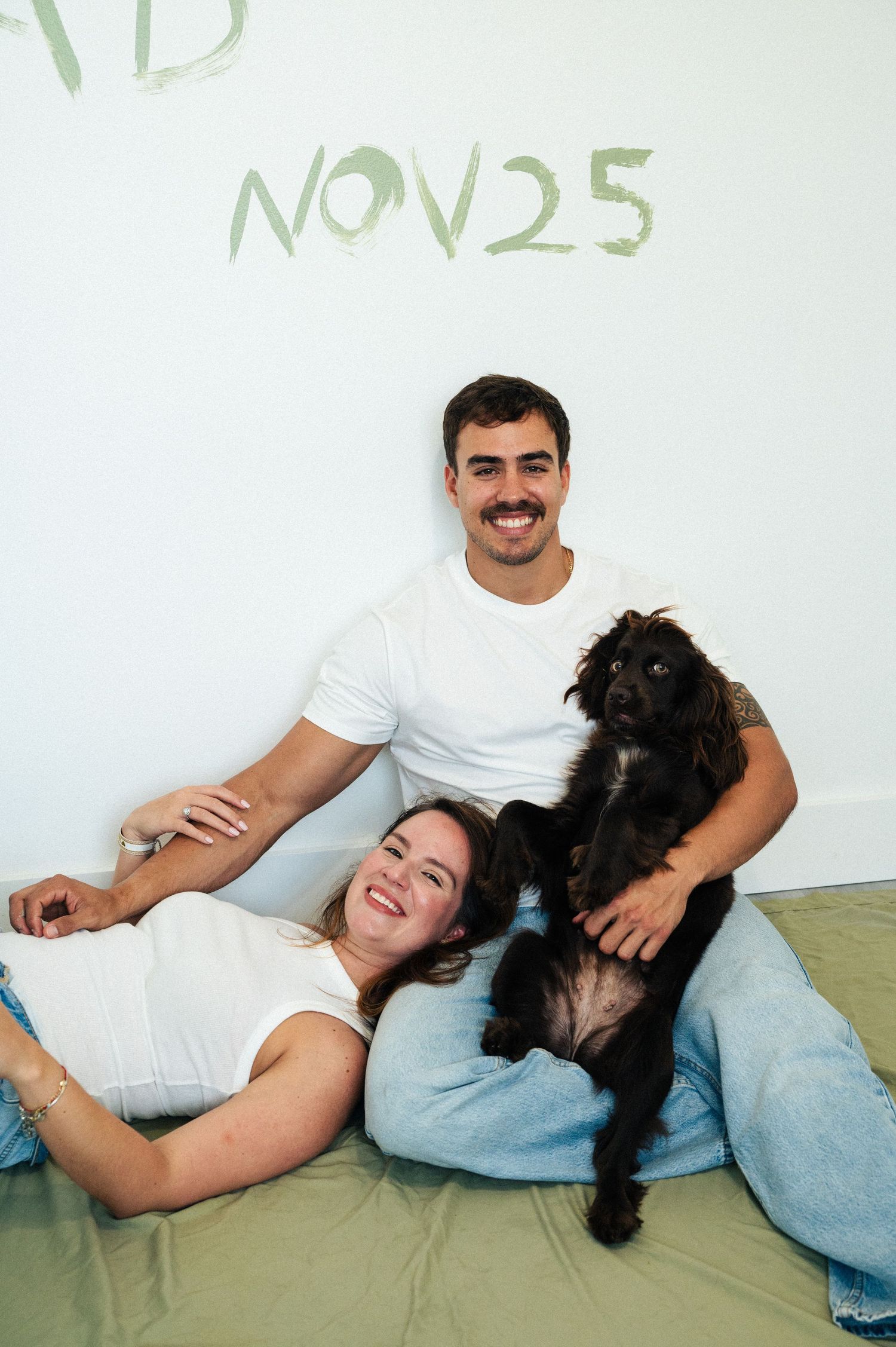 Happy couple relaxes on a green blanket with their black dog beneath a wall painted with the date NOV25.