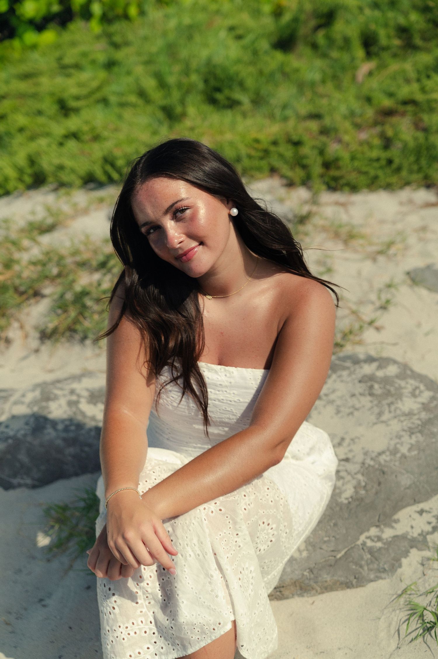 A smiling woman with long dark hair sits on a sandy rock in a white eyelet sundress, with green coastal vegetation behind her.