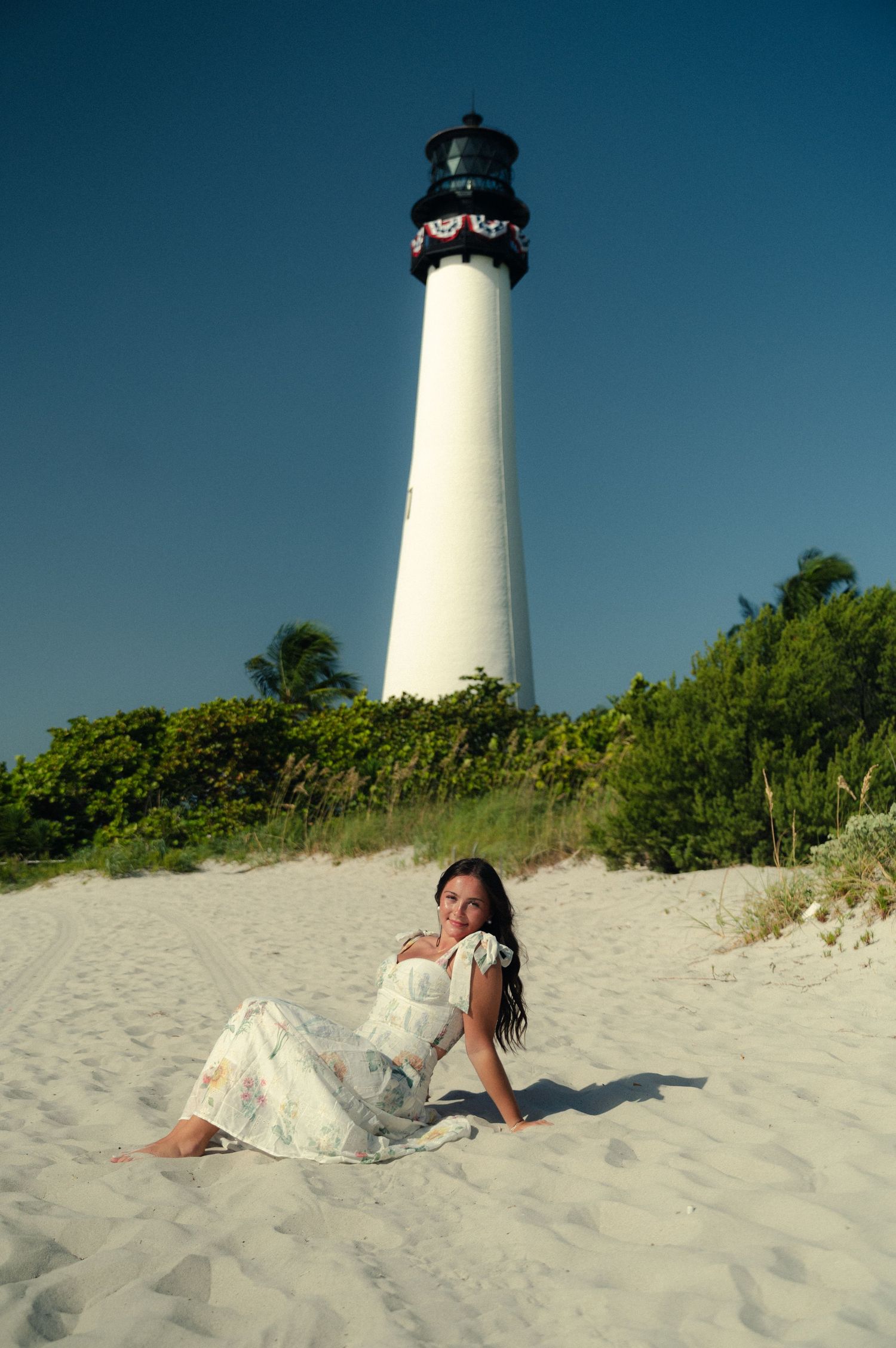 A woman in a flowing white dress reclines on a sandy beach beneath a tall white lighthouse and a clear blue sky.