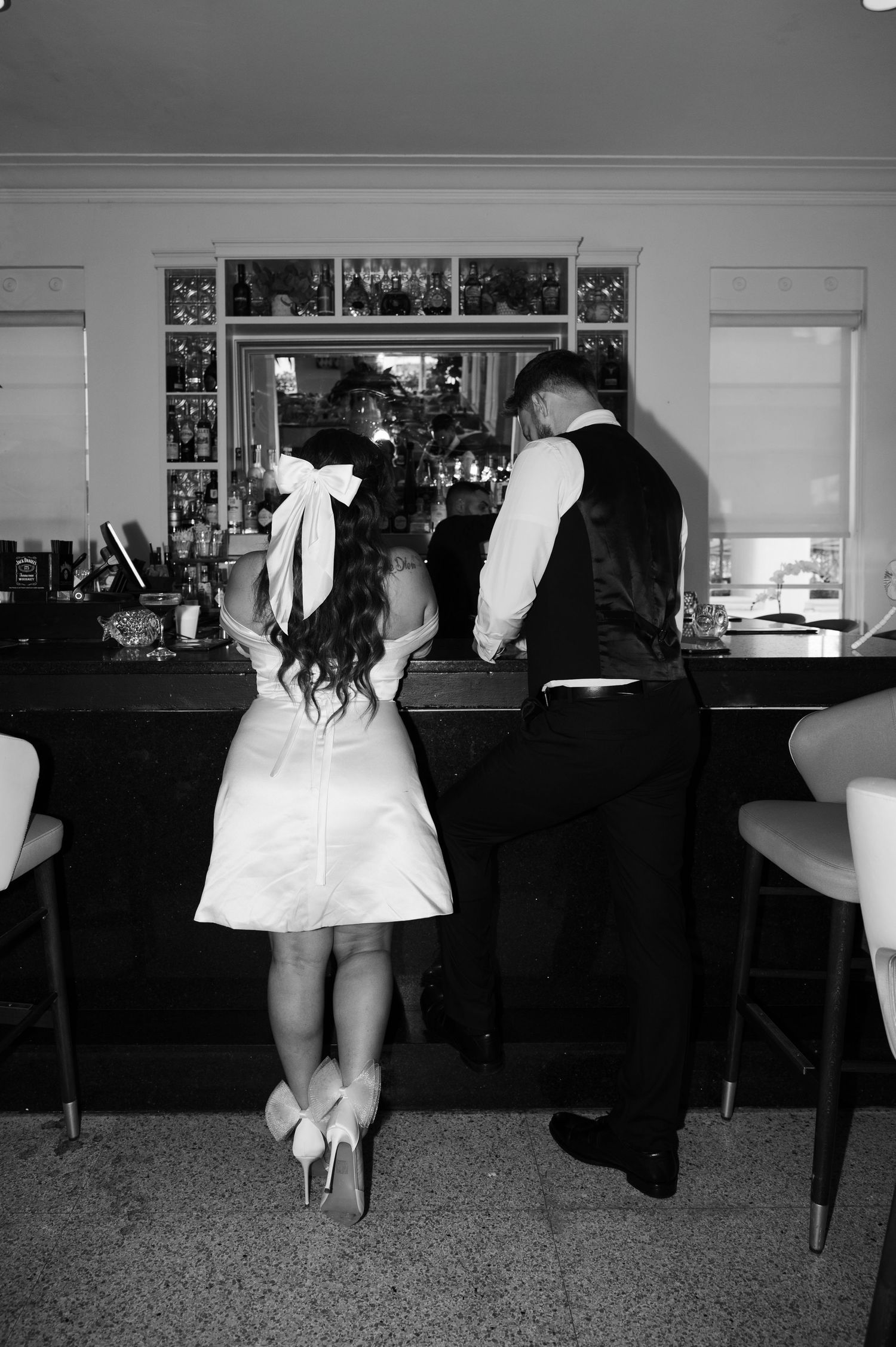 Black and white photo of a bride in a short dress with a large hair bow standing at a bar beside her groom.
