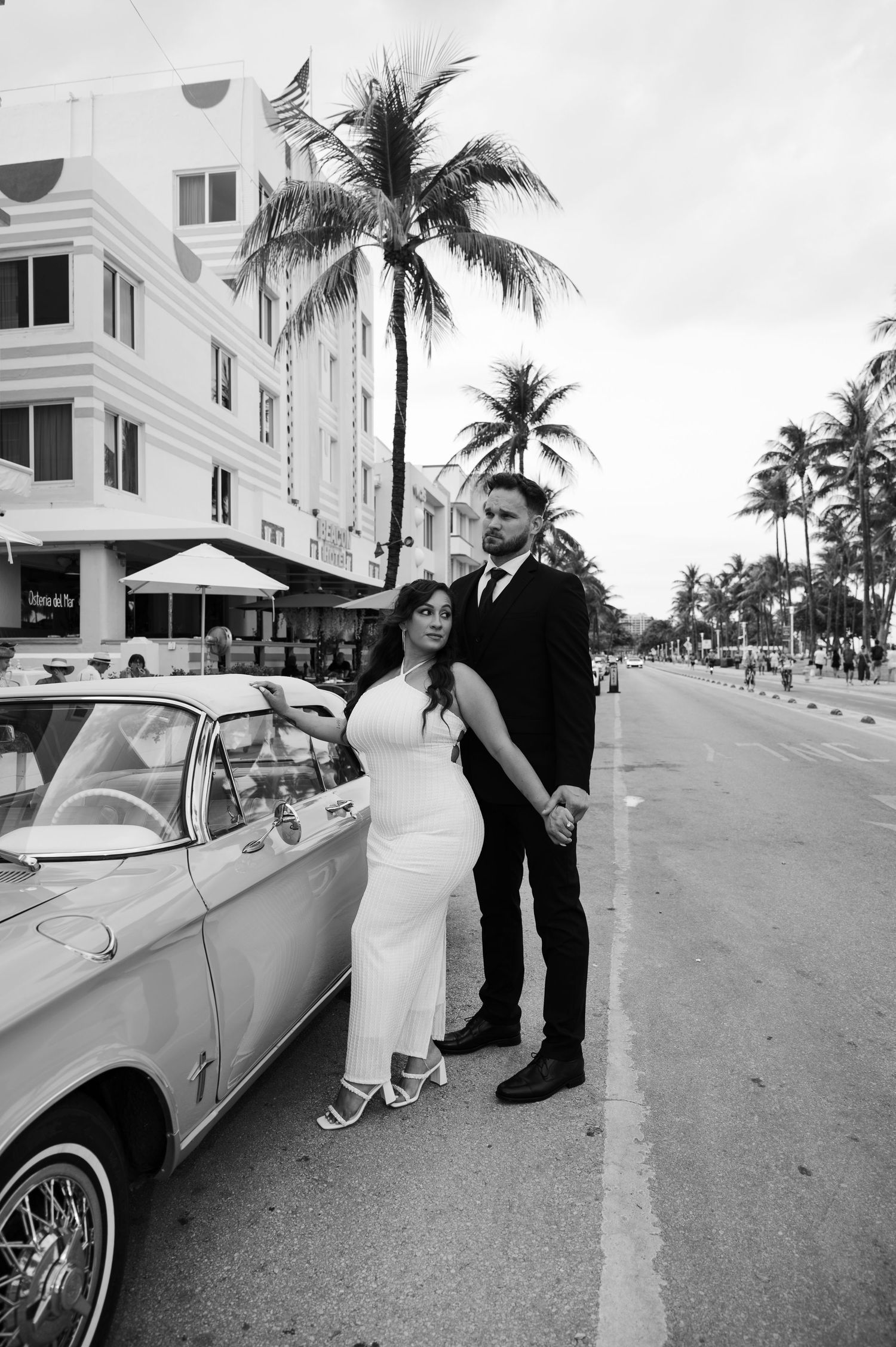 Black and white photo of a bride and groom posing beside a vintage car on a palm-tree-lined Miami street.