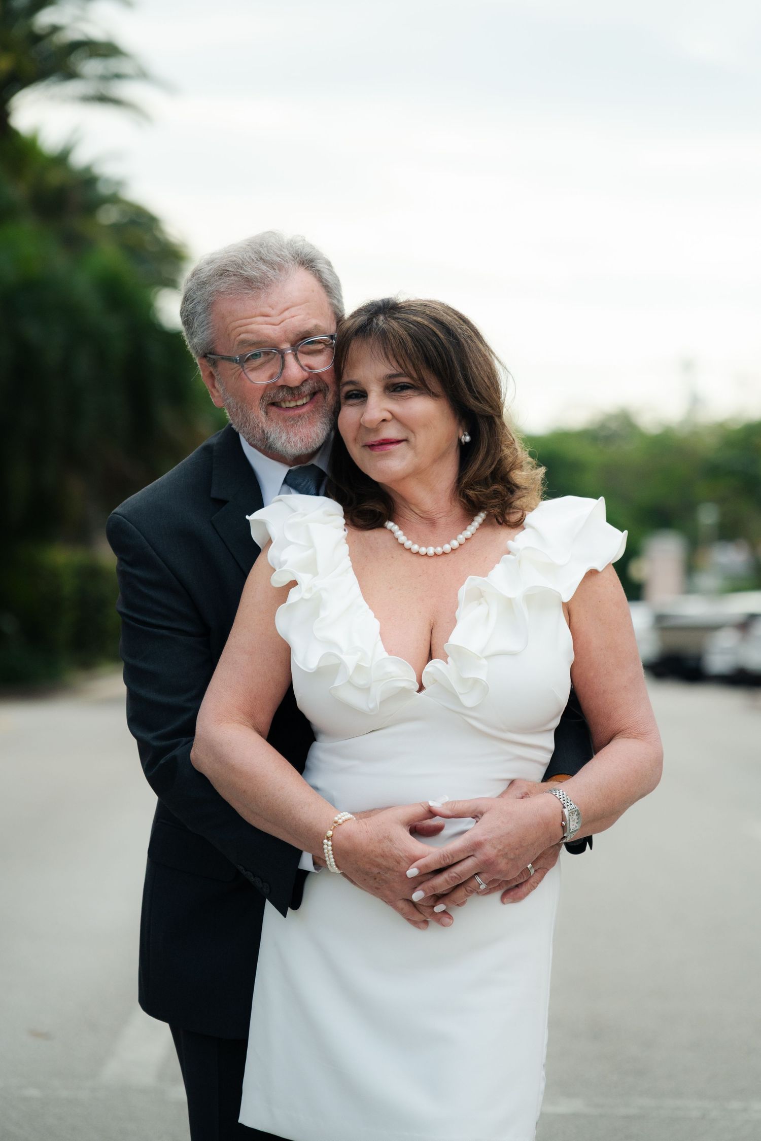 Smiling older man in a dark suit embraces a woman in a white ruffled dress and pearl necklace outdoors.