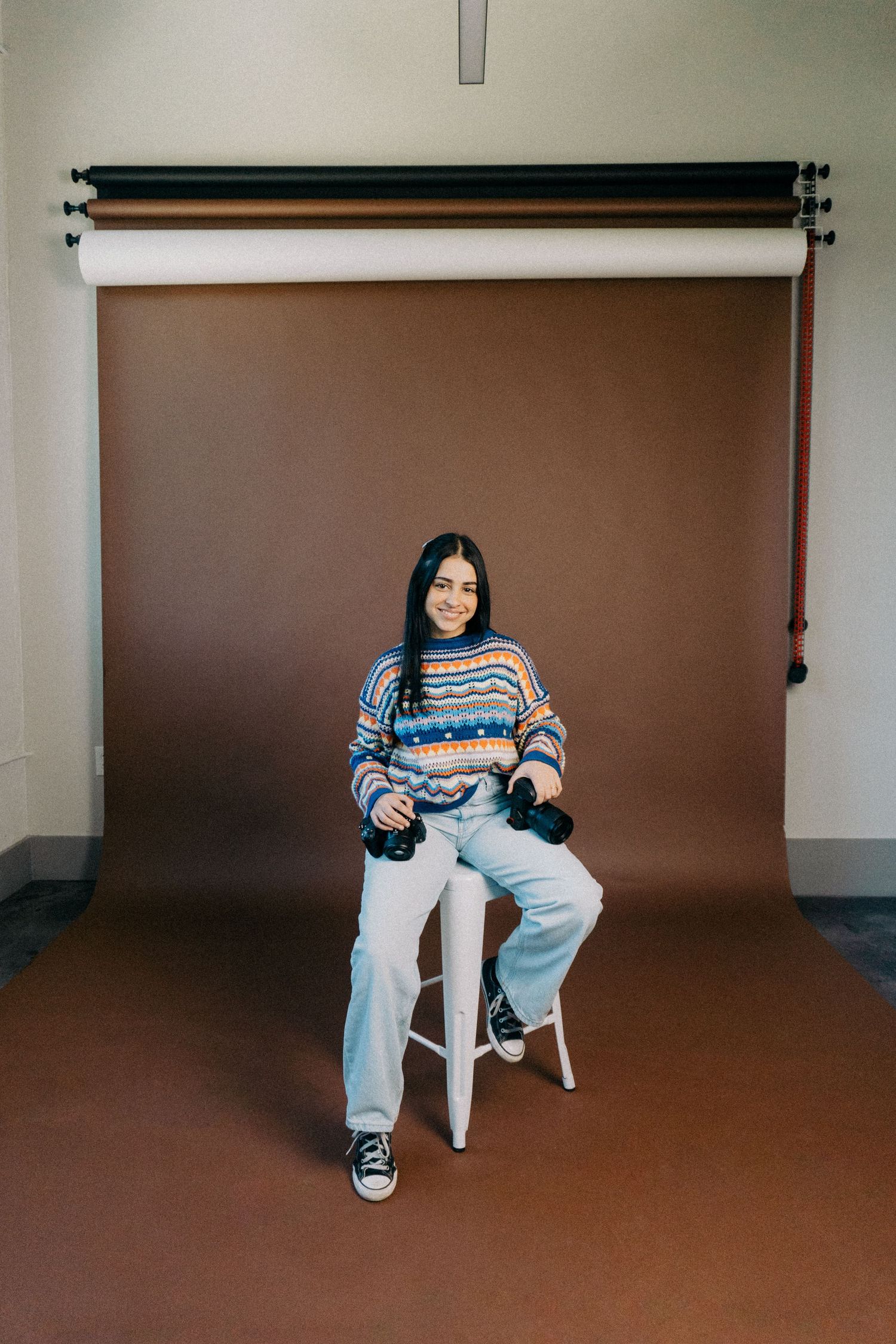 Photographer in a patterned sweater and jeans sitting on a stool holding cameras in front of a brown paper backdrop.
