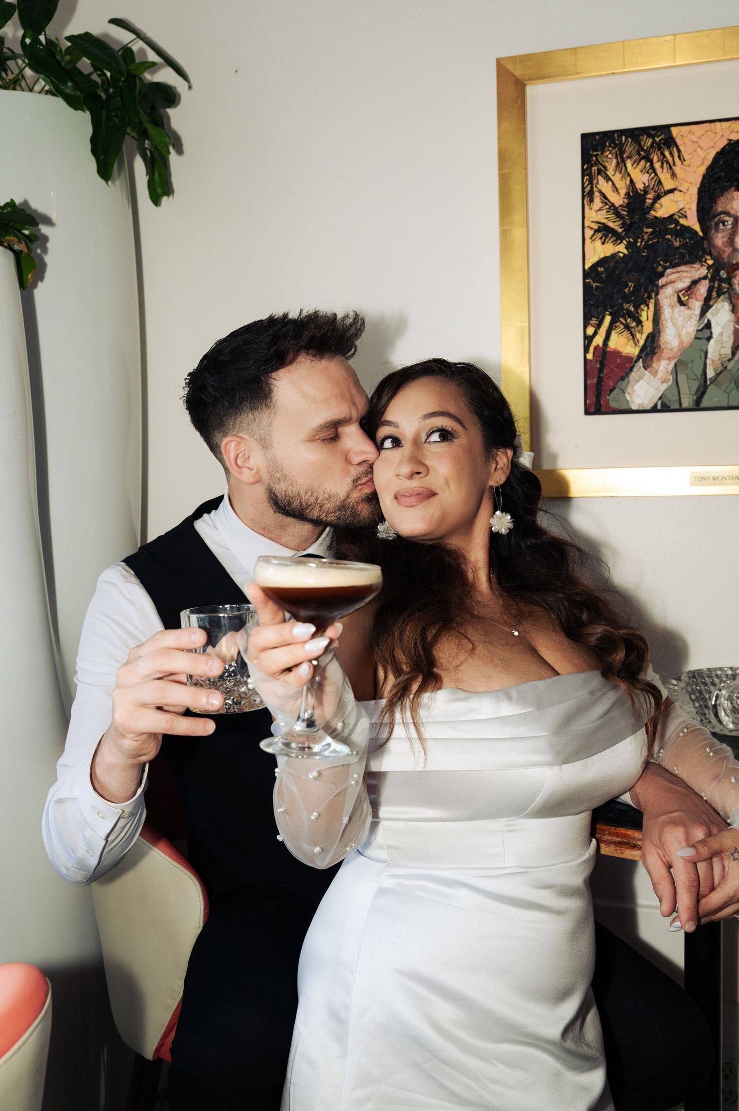 Groom kisses the cheek of a bride in a white satin dress as she holds an espresso martini cocktail.