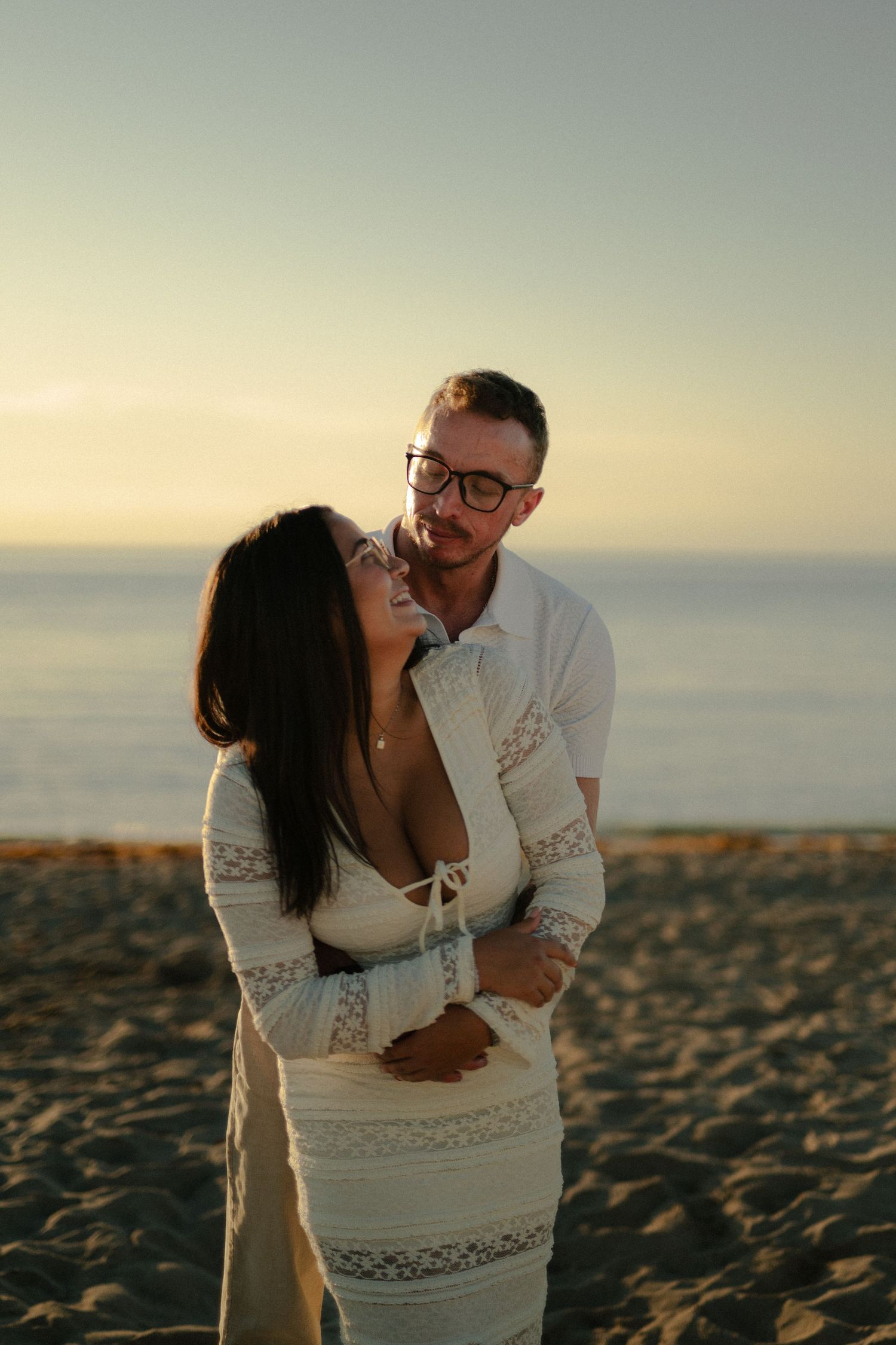 Couple in white embrace and laugh on the beach at golden hour, the man in glasses leaning in behind the woman in a lace dress.