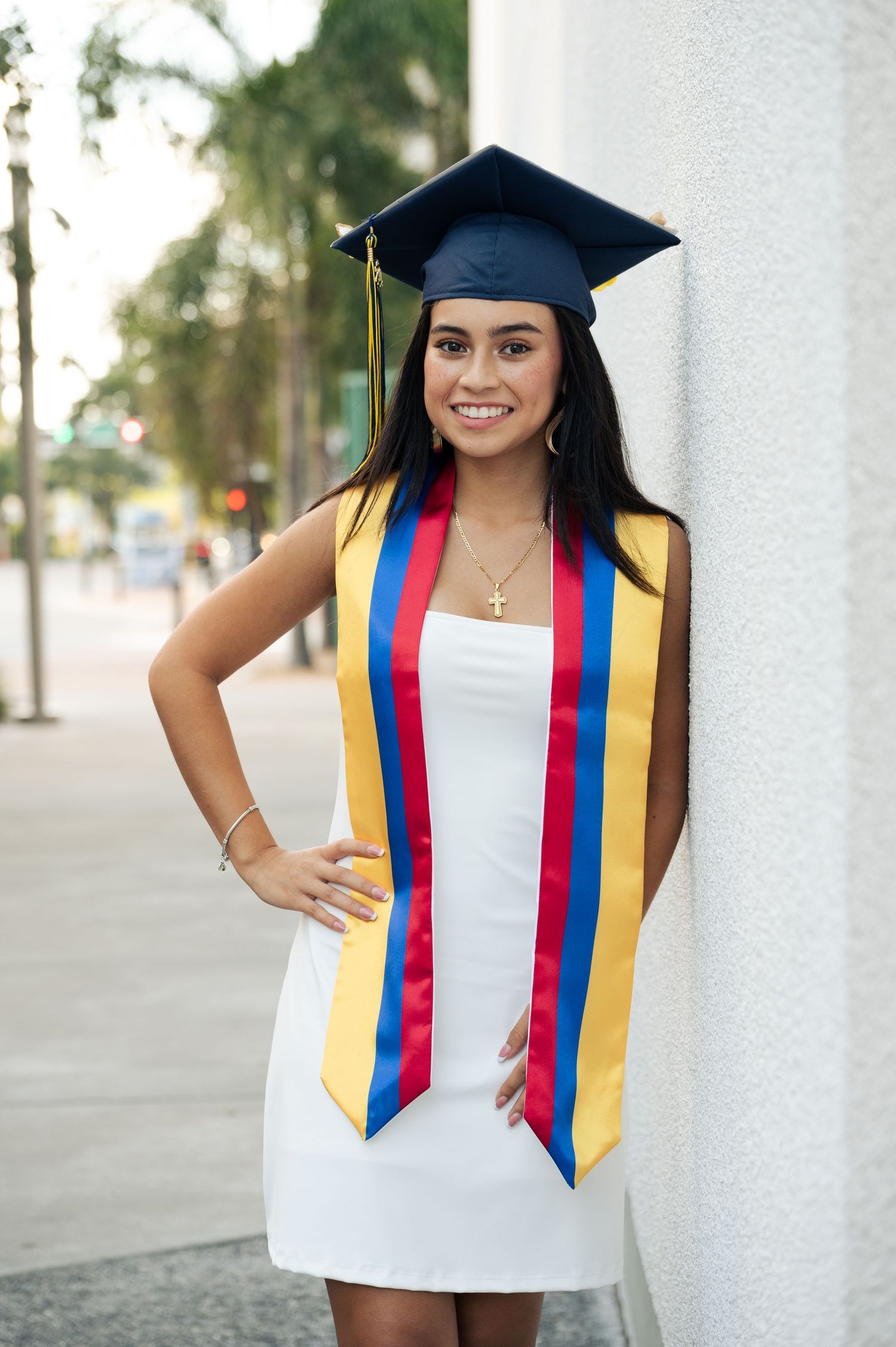 Smiling graduate in a navy cap and blue, red and yellow stole leans on a white wall in a white dress.