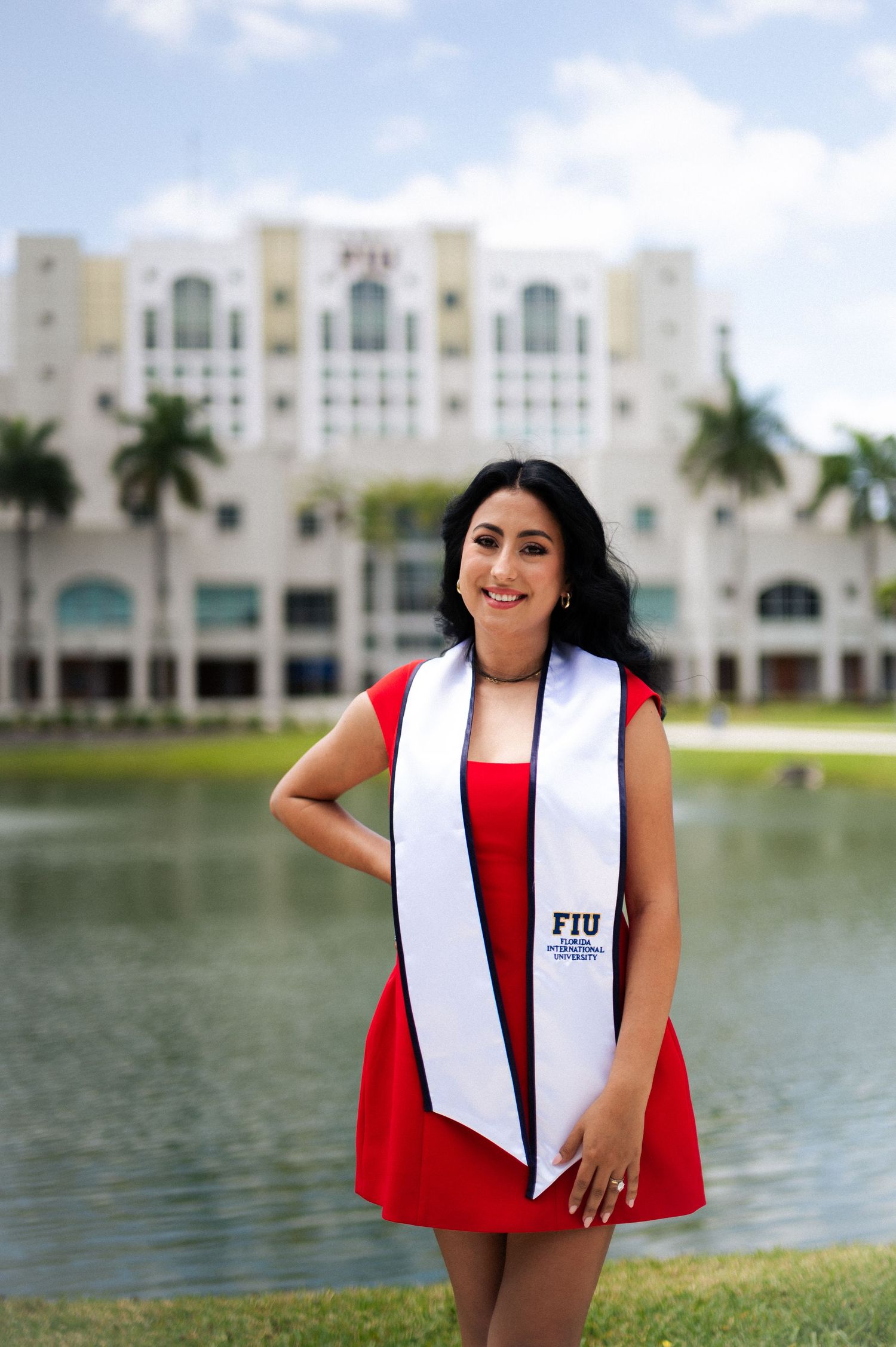 Smiling graduate in a red dress wearing an FIU graduation sash standing by a lake with a campus building behind her.