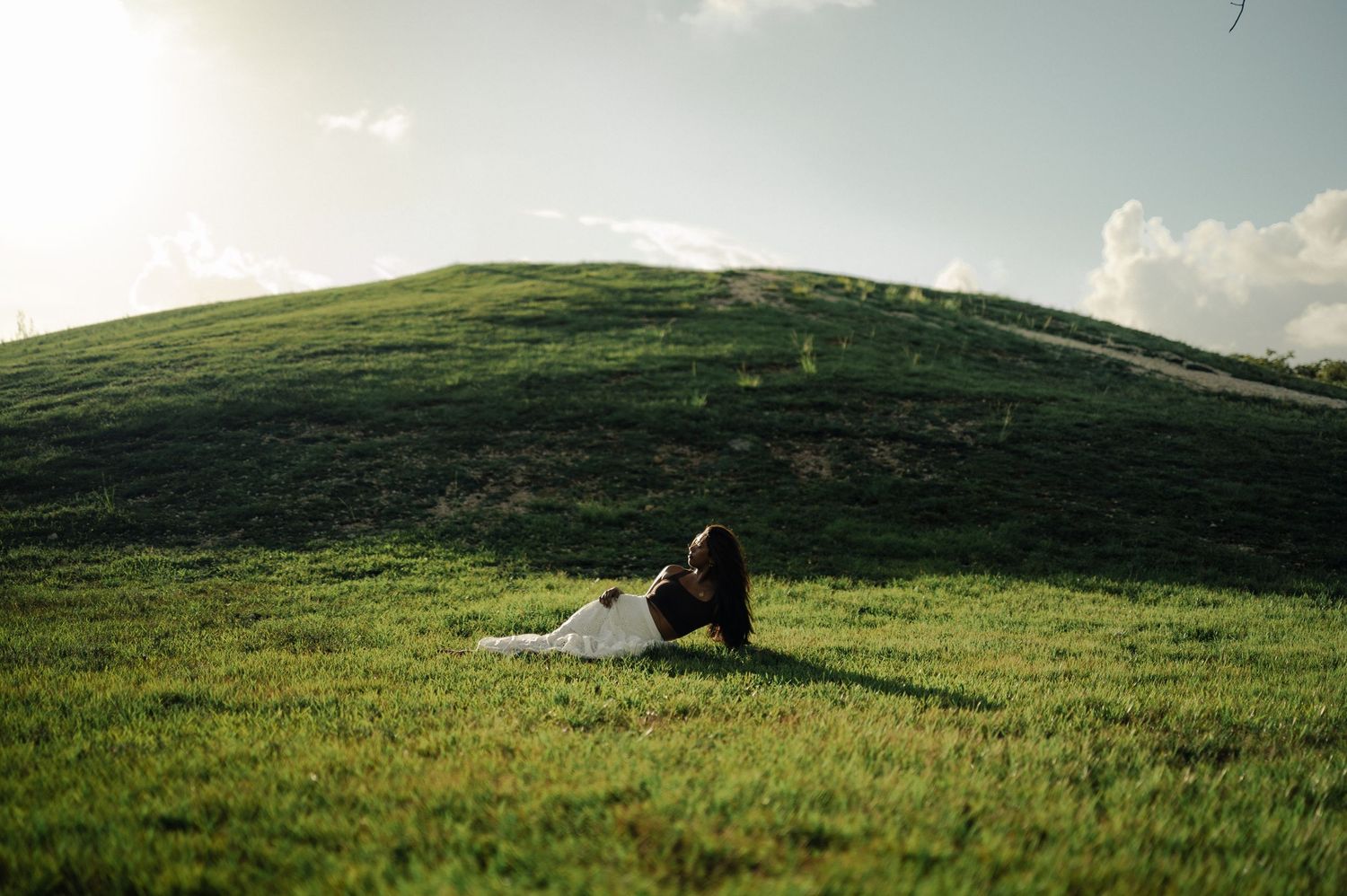 A woman in a white skirt rests on a grassy hillside in soft sunlight under a bright sky with scattered clouds.