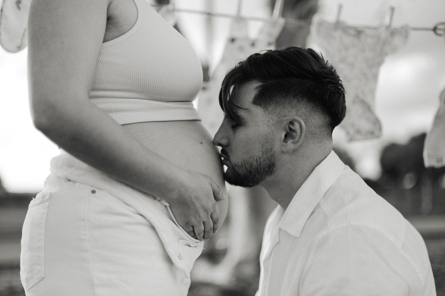 Black and white maternity photo of a man kissing his partner's pregnant belly, with baby clothes hanging behind them.