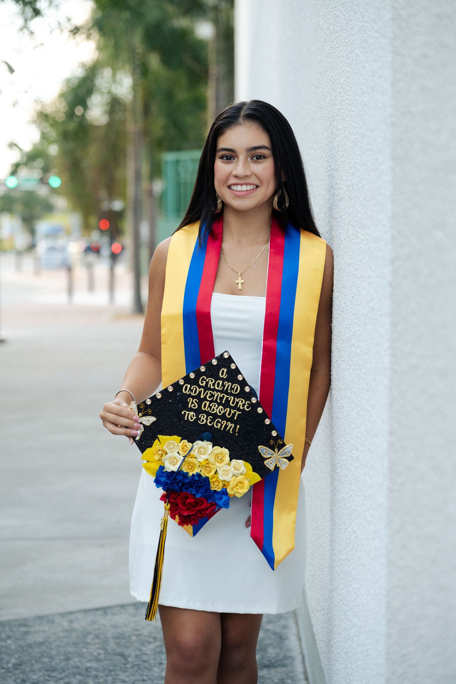 Smiling graduate in a white dress leans against a wall wearing a yellow, blue and red sash and a decorated grad cap.