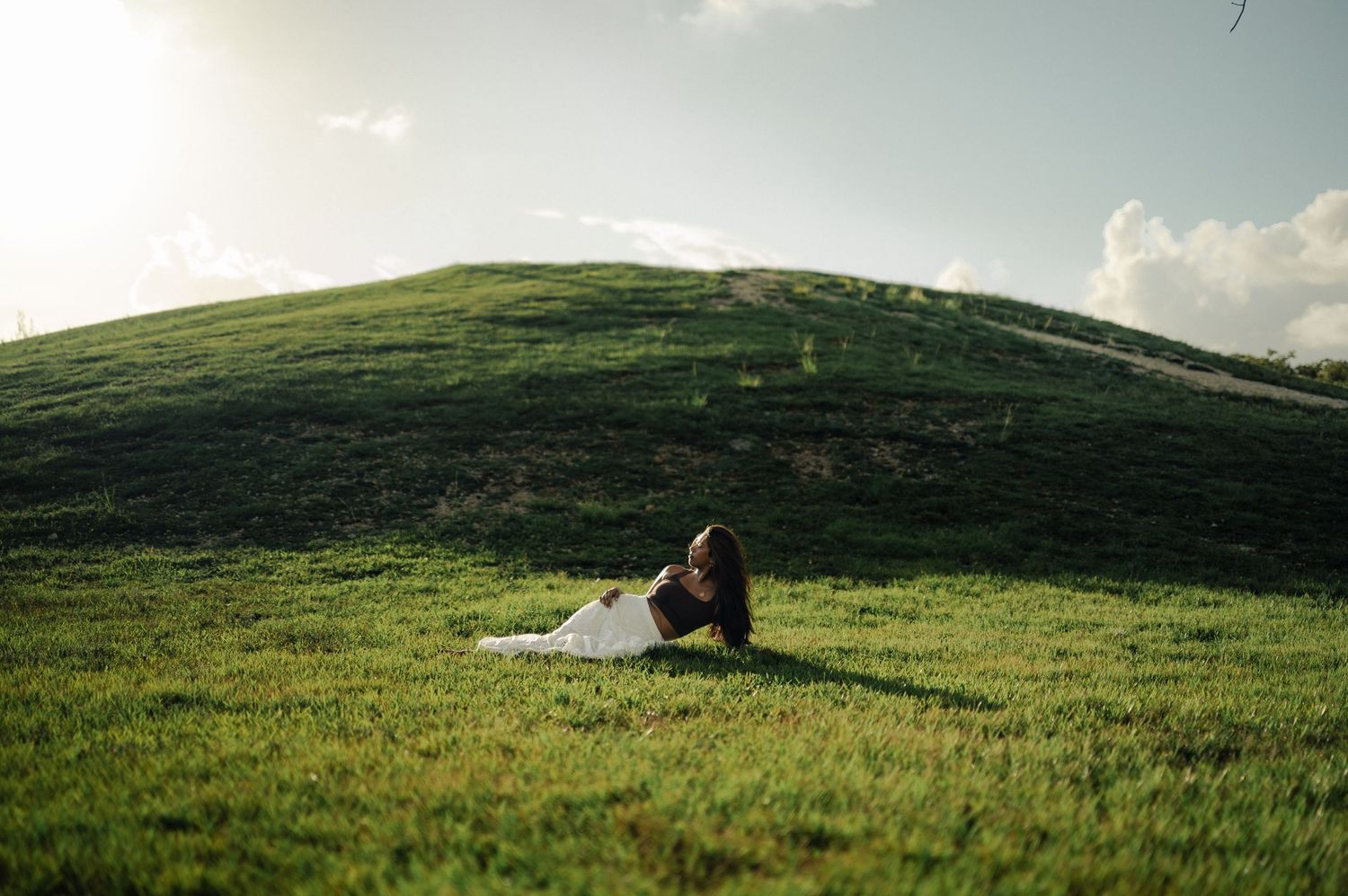 Woman in a white dress reclines on a green grassy hill under a bright sky with soft clouds.