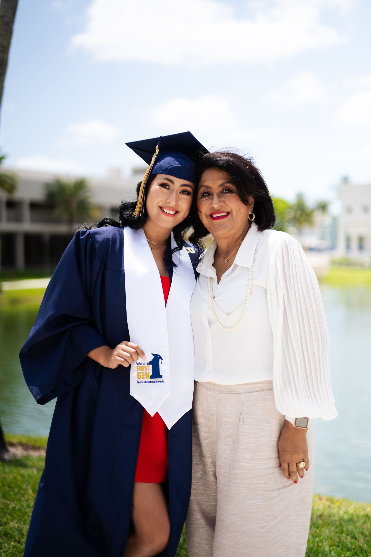 Smiling graduate in a navy gown and cap poses closely with an older woman in white beside a lake.