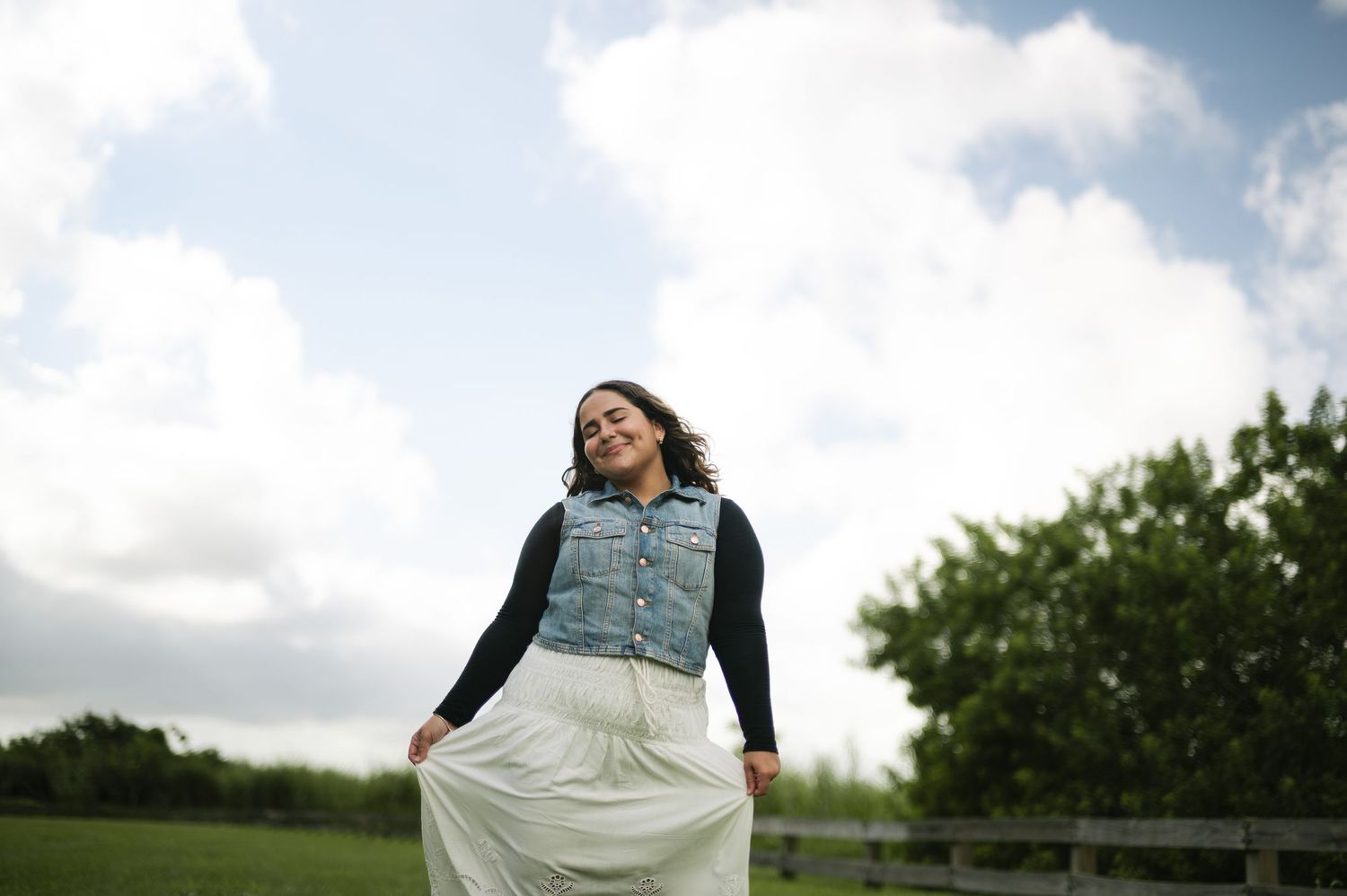 Smiling woman in a denim vest and white skirt twirling in a green field under a cloudy sky.