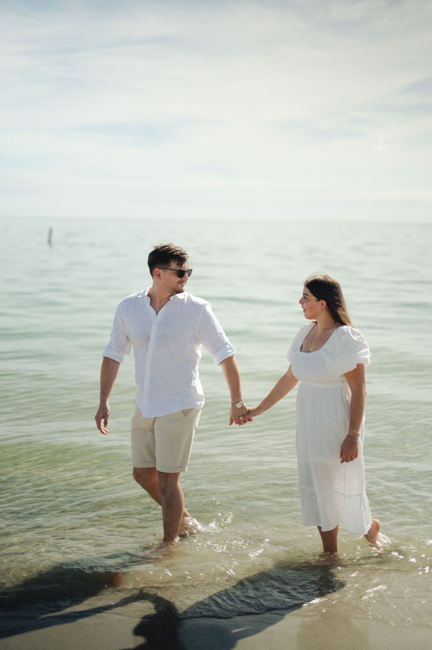 Expecting couple in white outfits hold hands and wade through shallow turquoise water at the beach.