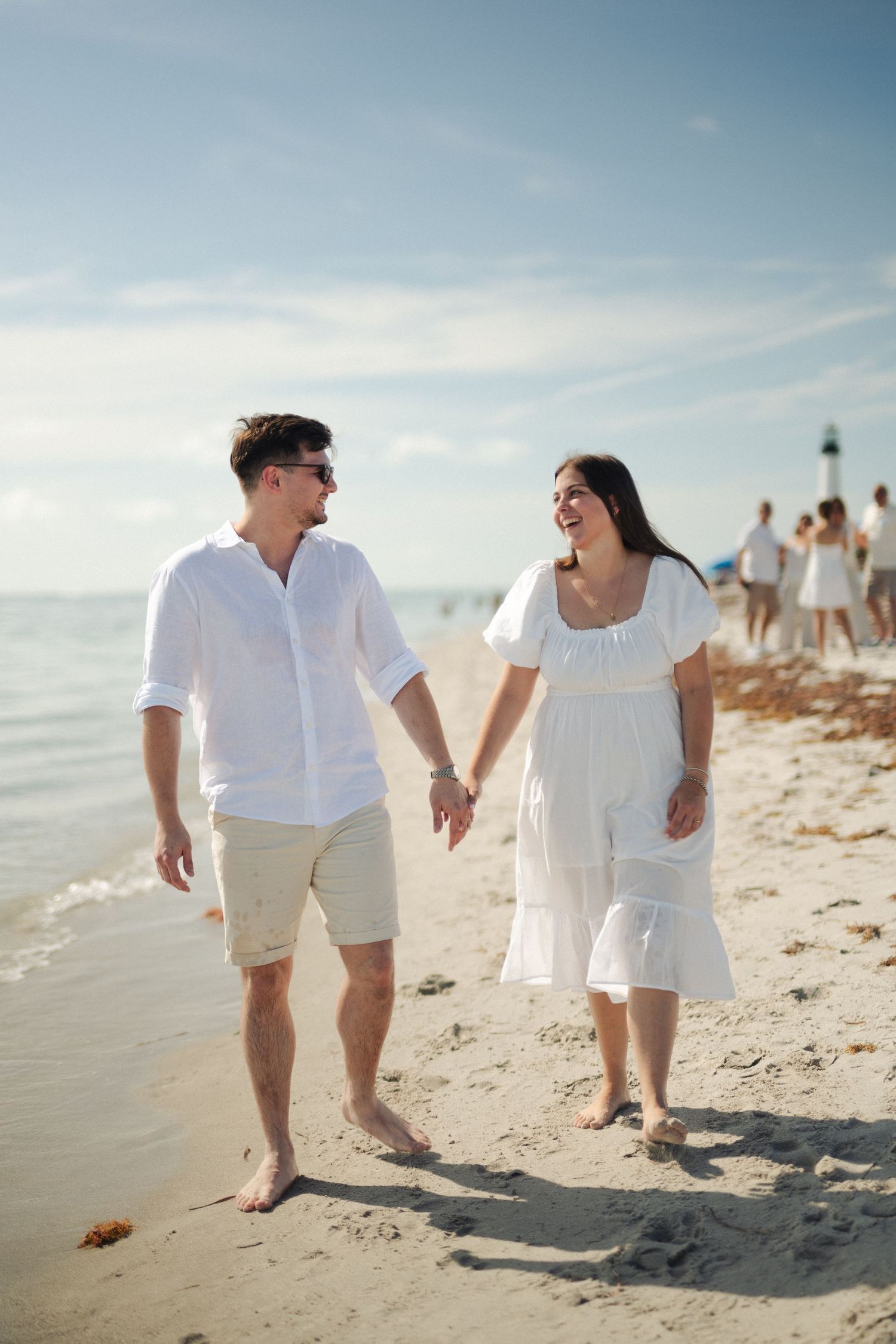 Smiling couple in white outfits holding hands and walking barefoot along a sandy beach near a lighthouse.