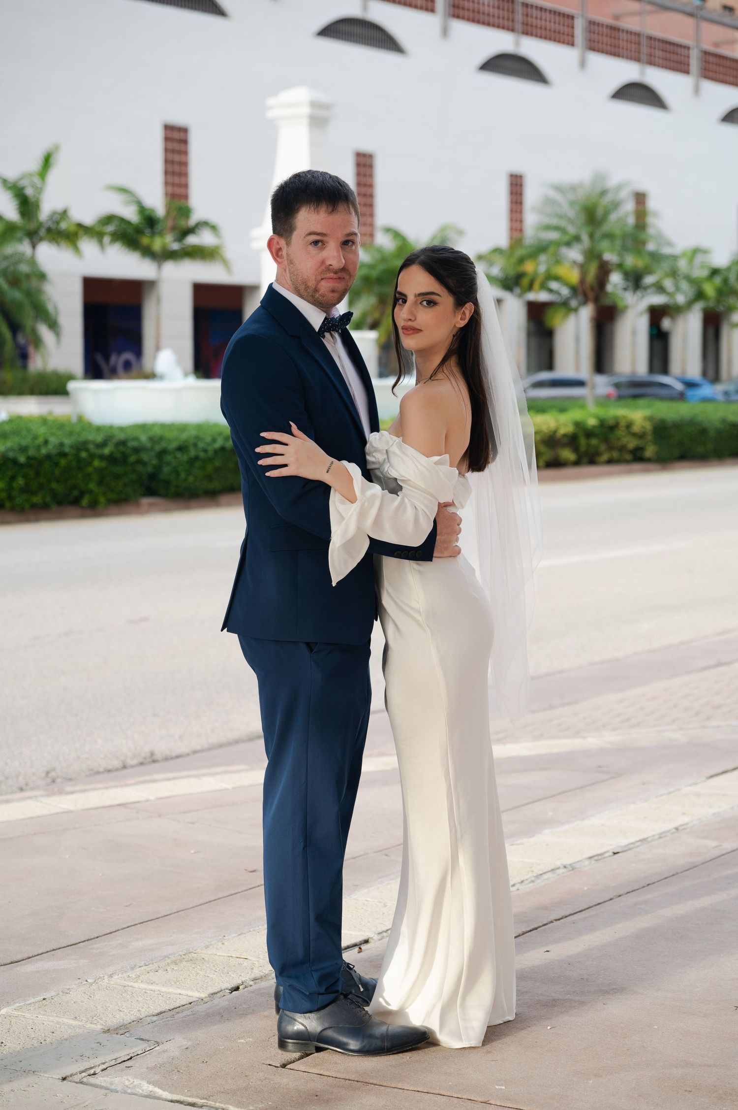 Bride in a white gown and veil embraces her groom in a navy suit on a palm-lined street during a sunny wedding photo session.
