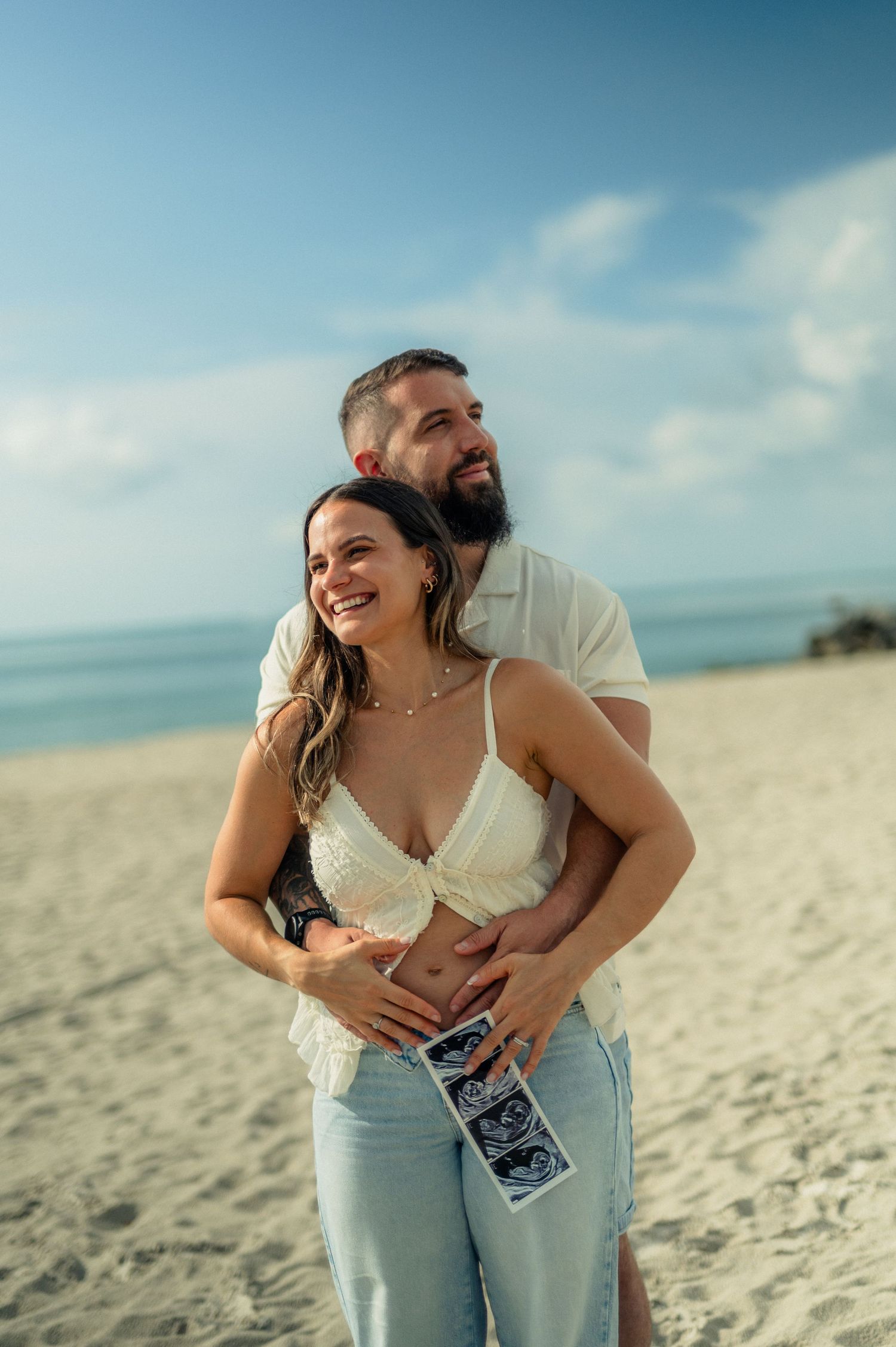 Smiling woman holds ultrasound photos at her belly as her bearded partner hugs her from behind on a sunny beach.