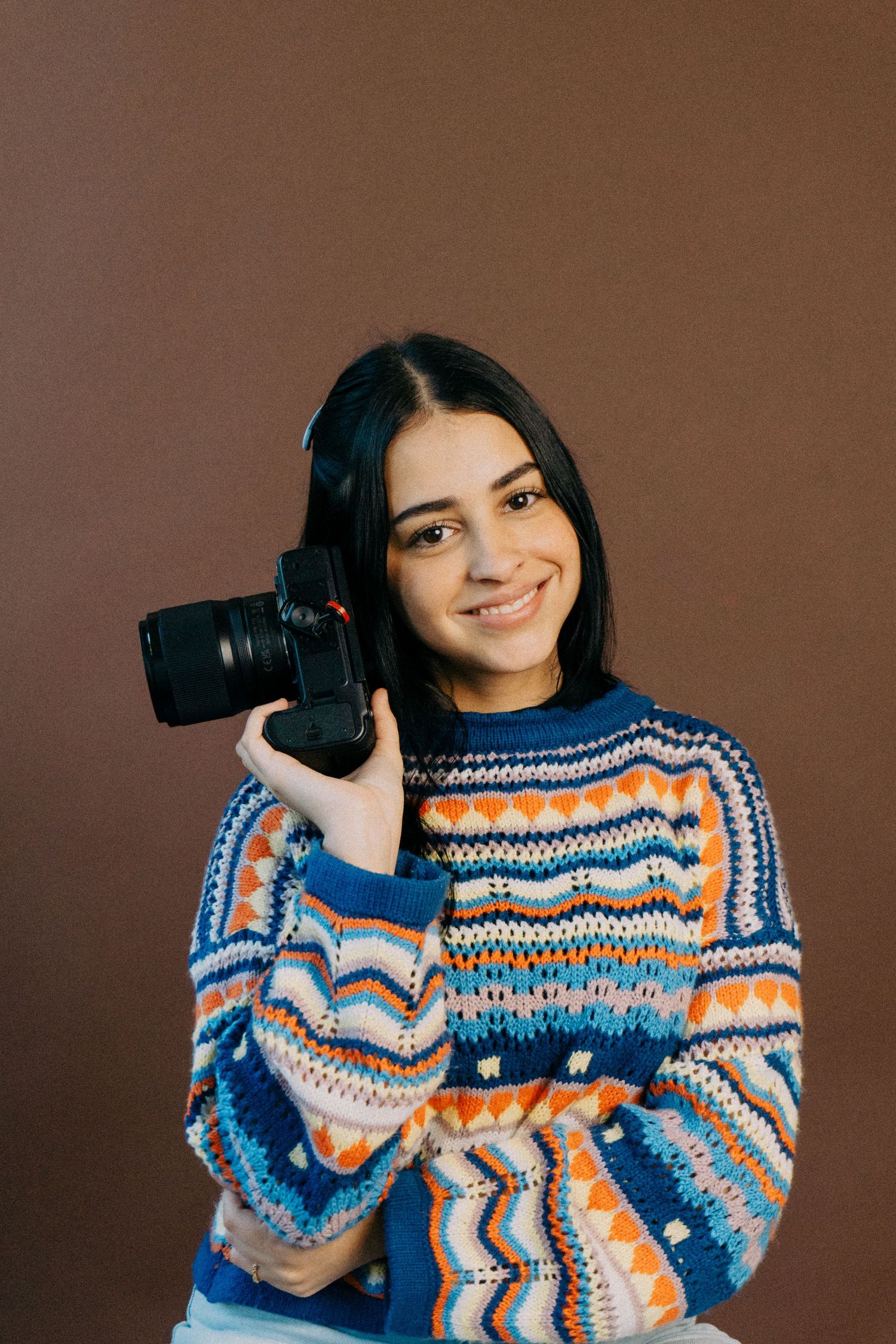 Cheerful young woman in a blue and orange knit sweater holds a black camera beside her face in front of a brown backdrop.