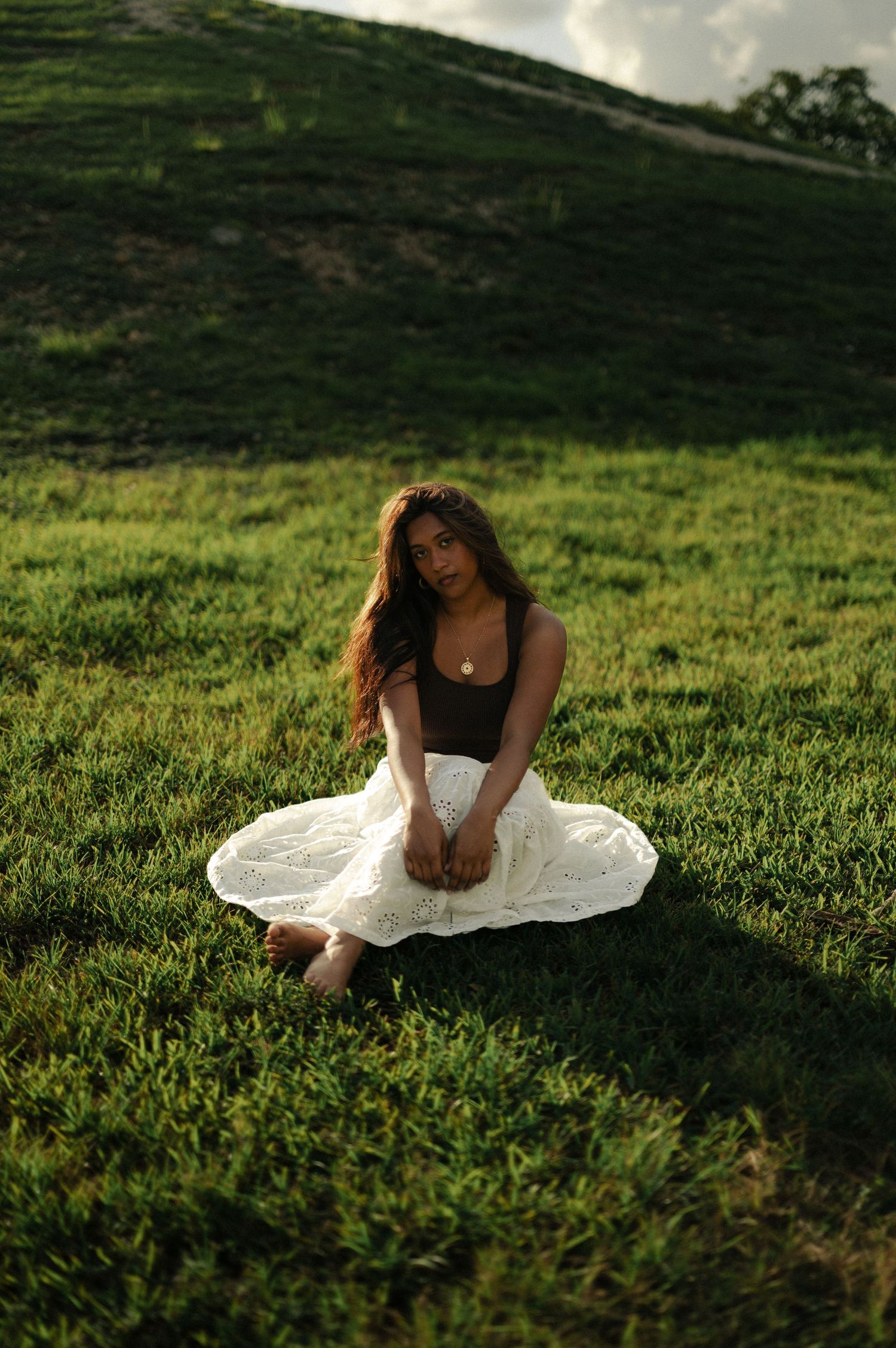 Woman in a white skirt and dark top sits cross-legged on green grass in warm golden sunlight.
