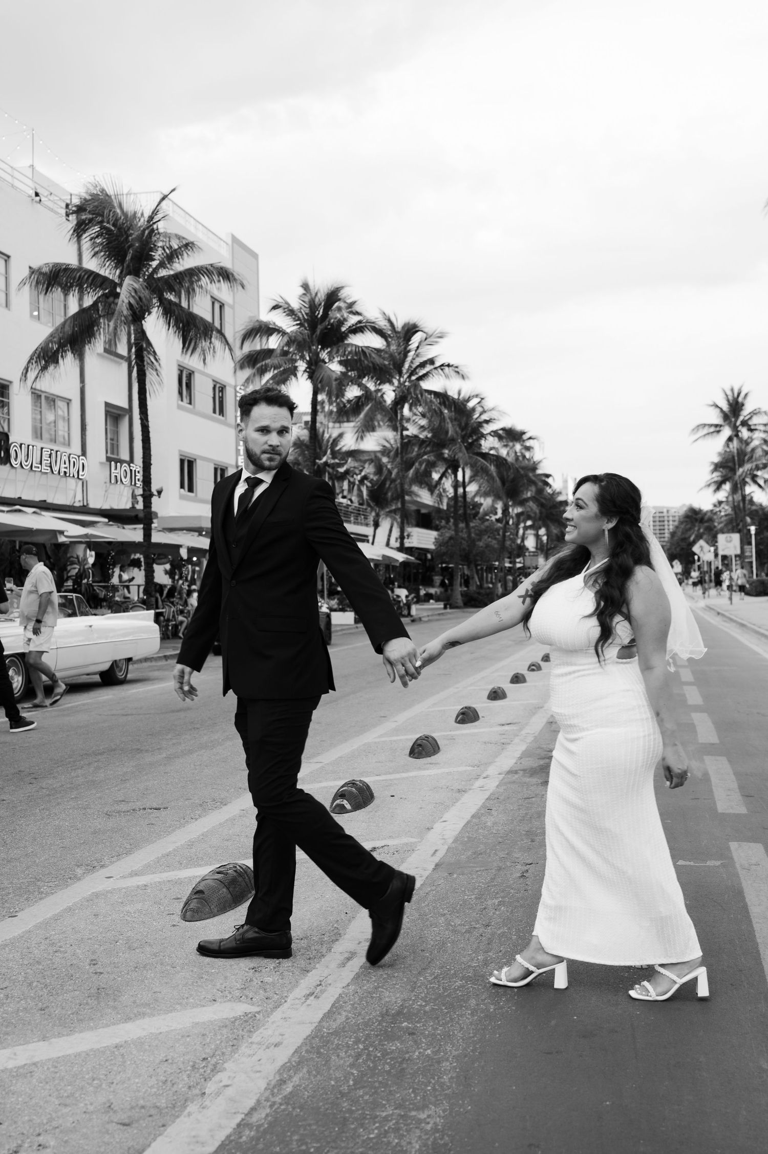 Black and white photo of a bride and groom holding hands while crossing a Miami Beach street lined with palm trees.