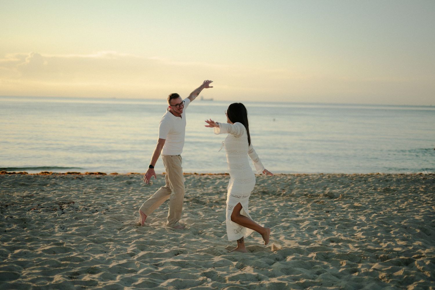 Couple in white outfits dance joyfully with open arms on a sandy beach at dusk beside the calm ocean during an engagement shoot.