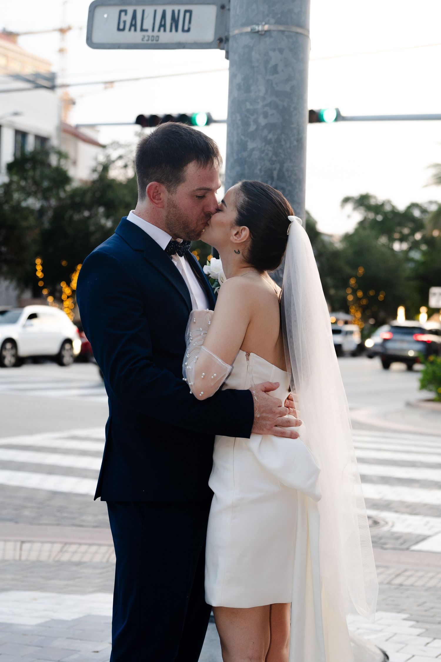 Groom in a navy suit kisses his bride in a short white dress and veil at a Galiano street corner during their wedding photos.