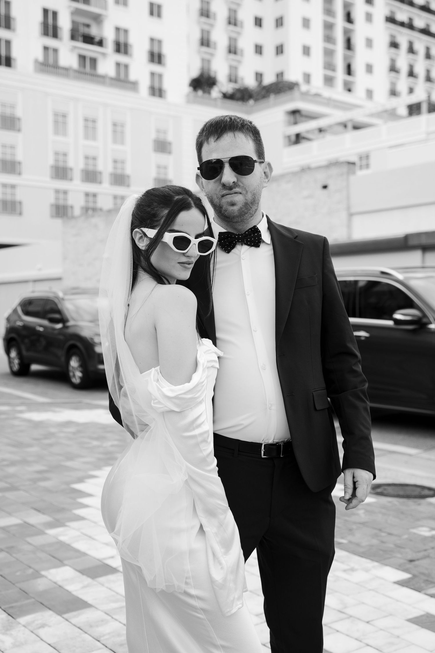 Black and white portrait of a bride in white sunglasses and a groom in a bow tie posing on a city street after their wedding.