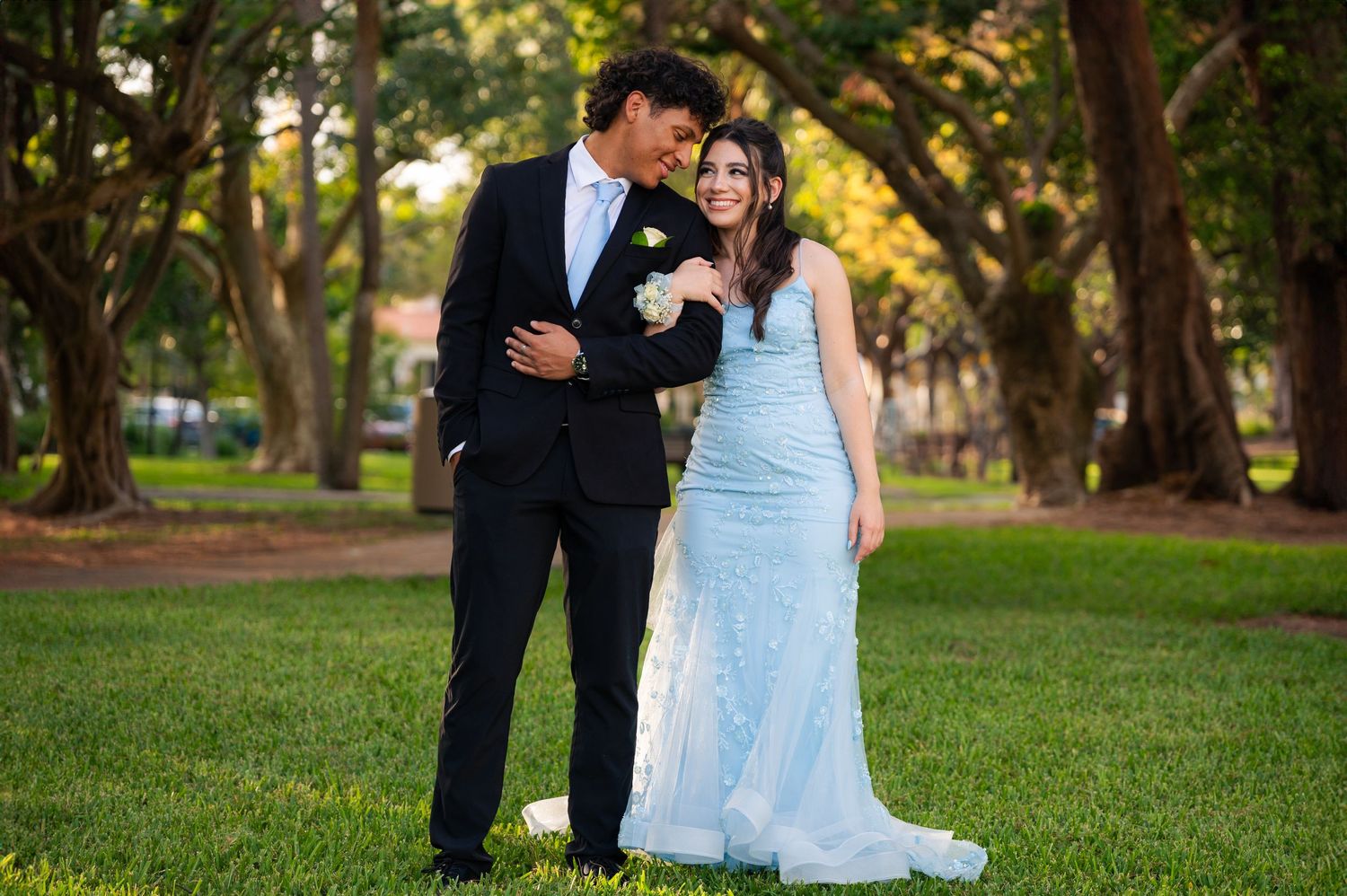 A smiling couple in formal wear, he in a black suit with a boutonniere and she in a light blue gown, pose in a leafy park.