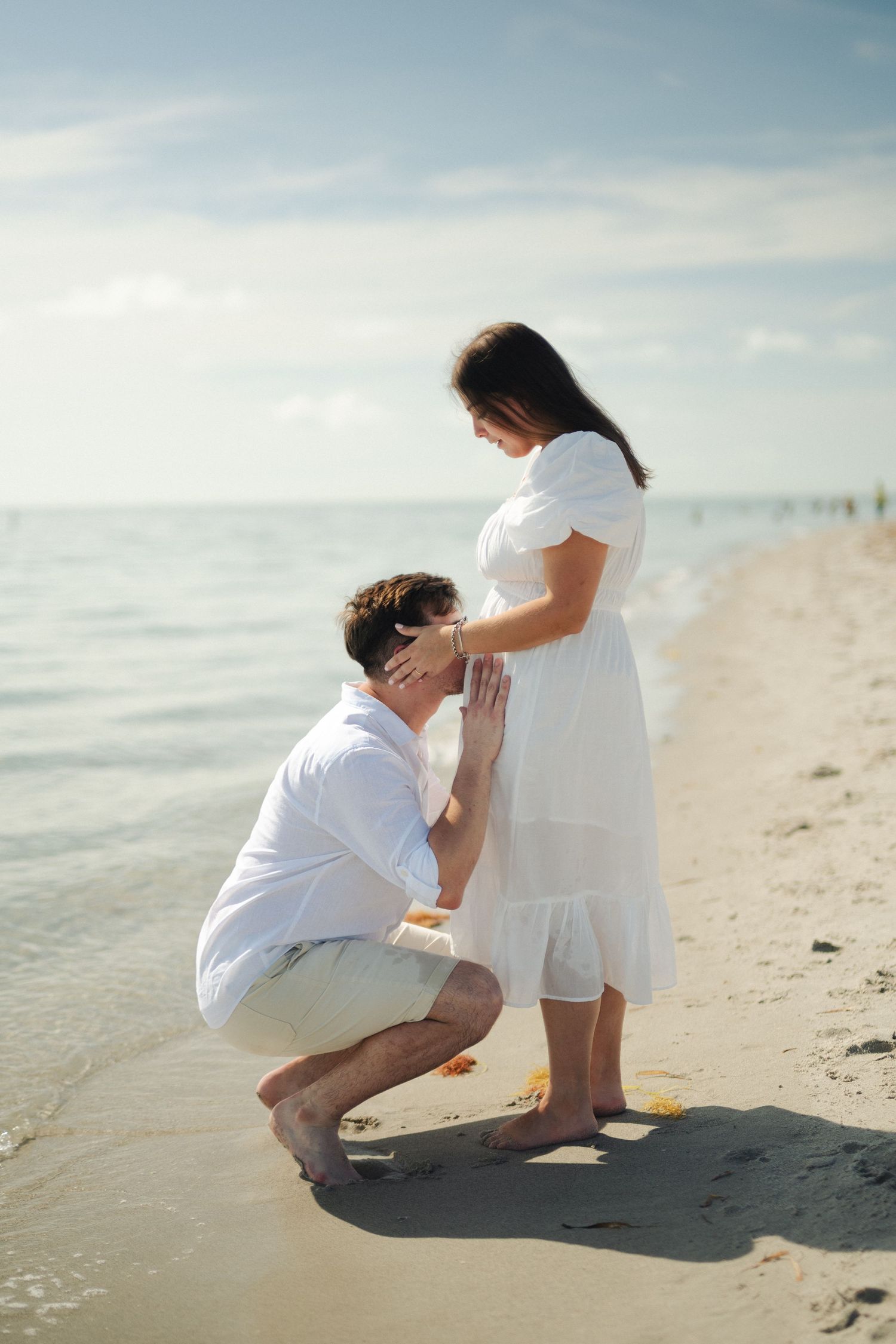 Man in white shirt kneels on the sandy beach and kisses the baby bump of his pregnant partner in a white dress.