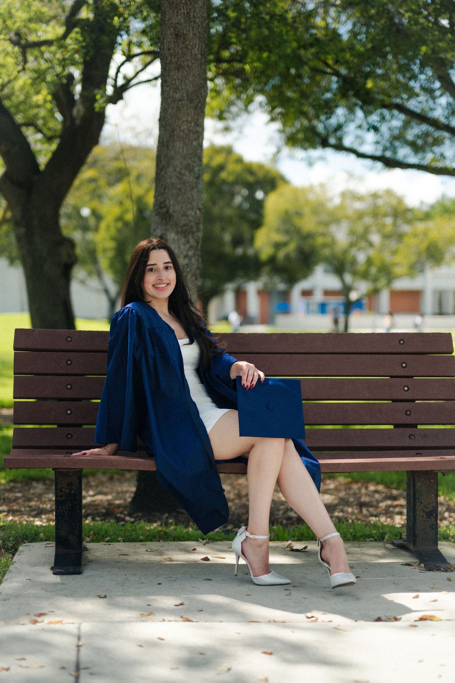 Graduate in a navy gown and white heels sits on a park bench under a large tree on a sunny campus.