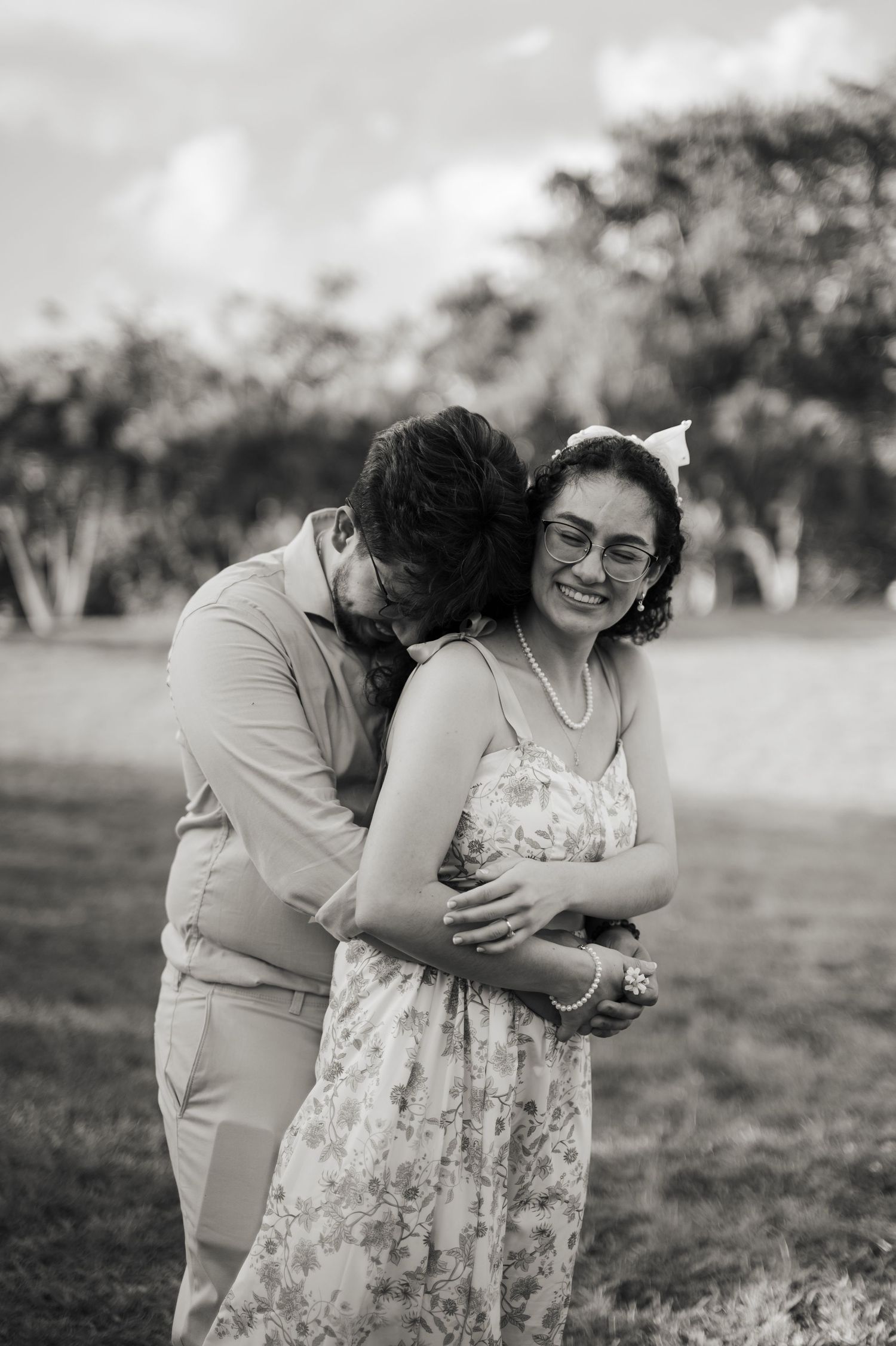 Black and white photo of a man hugging a smiling woman in a floral dress outdoors.