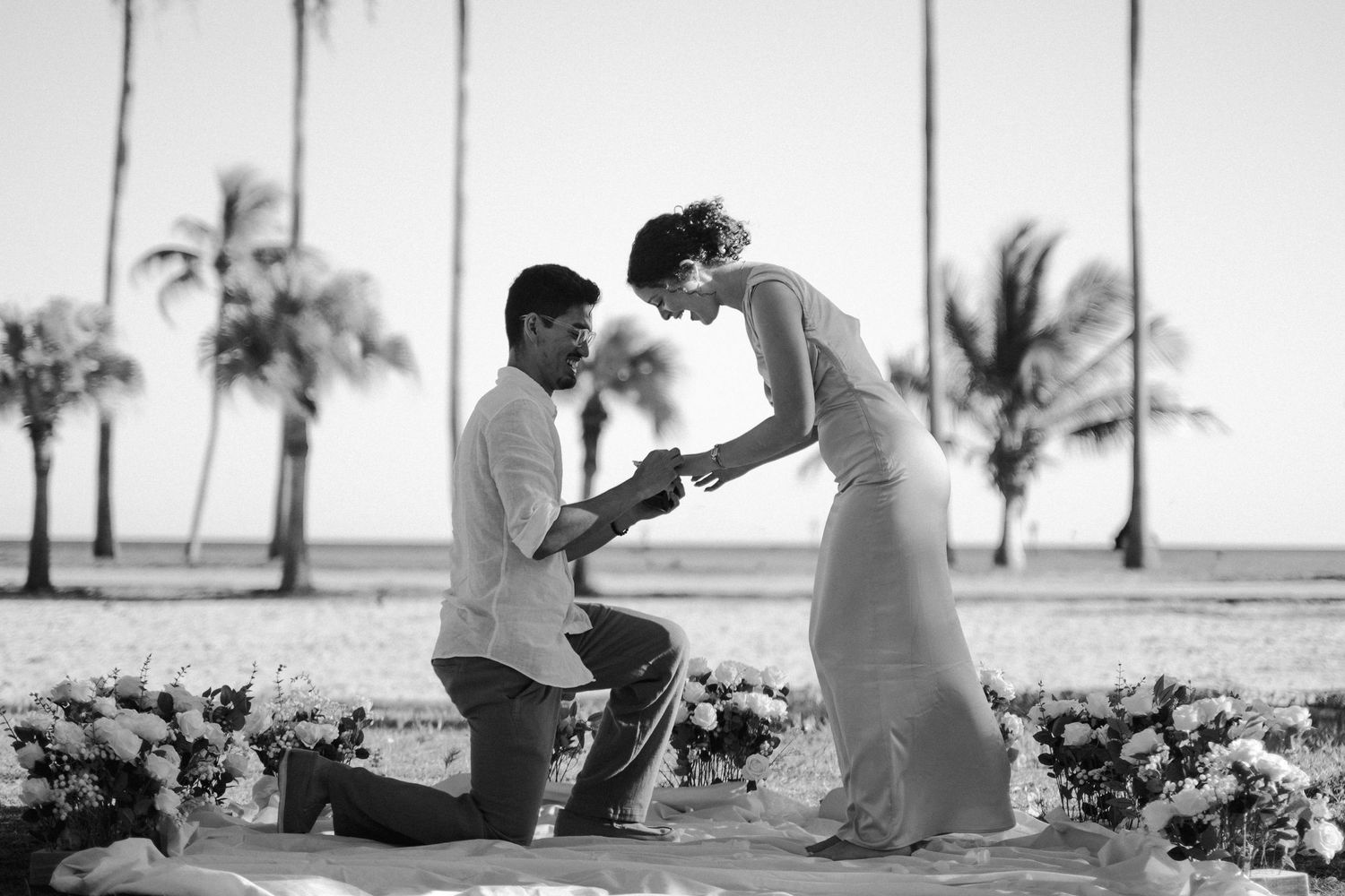 A man kneels on a sandy beach among flowers, proposing to a woman in a long dress, with palm trees behind them.