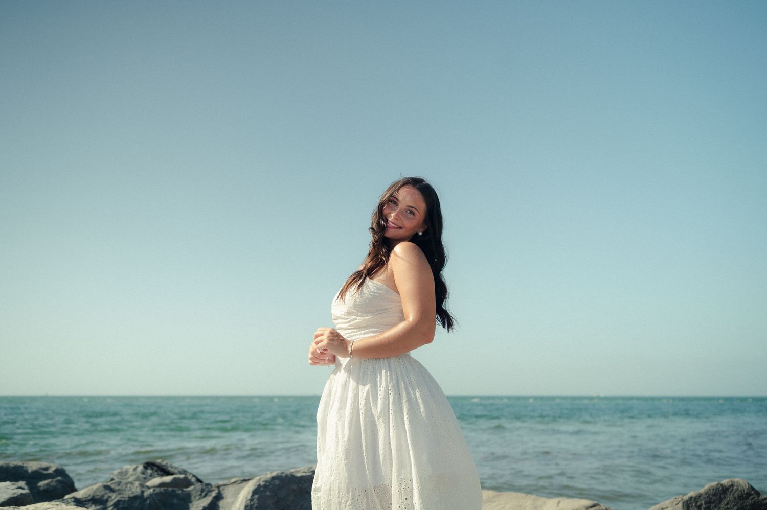 A woman in a white sundress stands on rocks by the ocean, gazing at the camera under a pale blue sky.