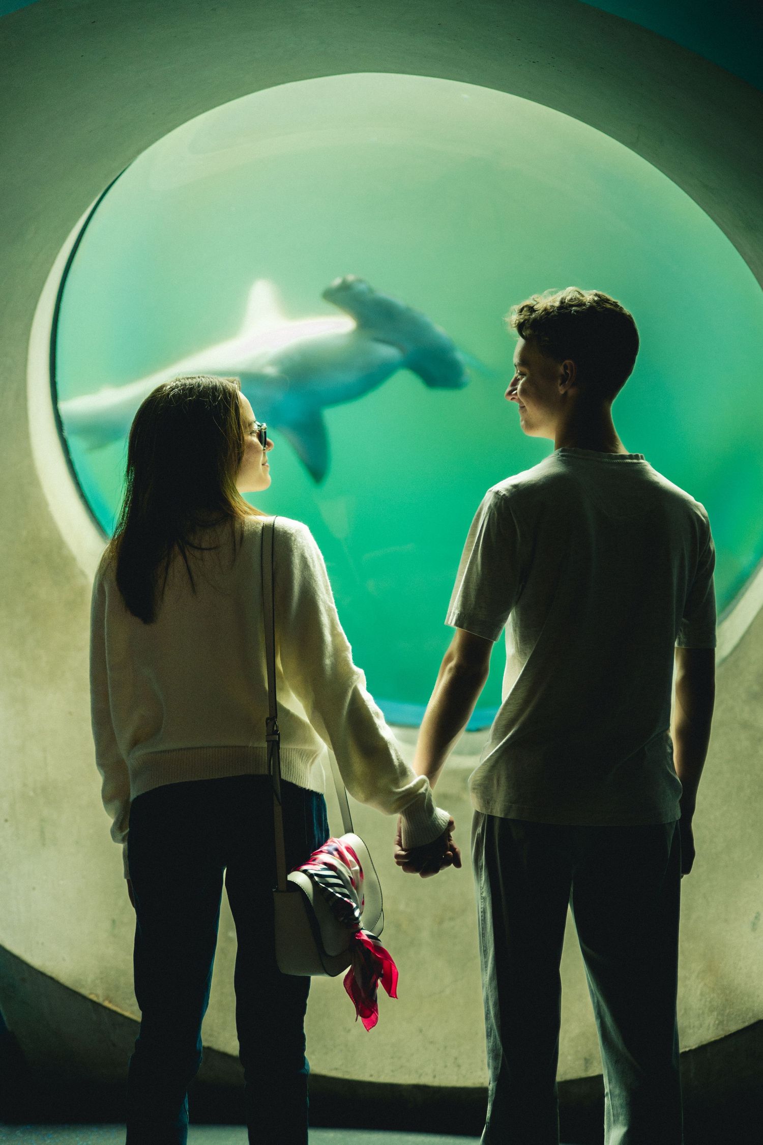 Couple holds hands in front of a large round aquarium window as a shark swims through glowing green water.