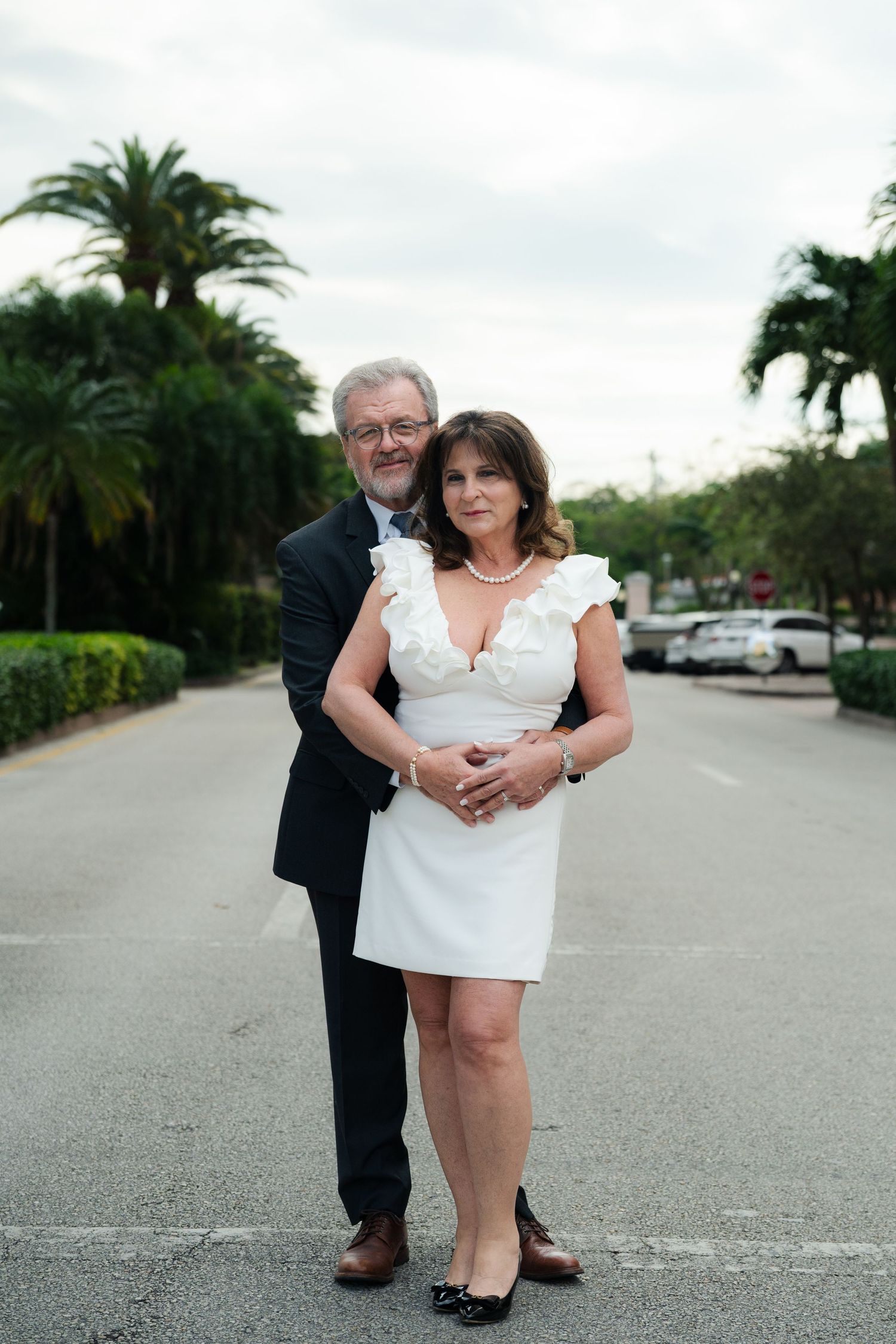 Mature couple embraces on a quiet palm-lined street, the woman in a white ruffled dress and pearls and the man in a dark suit.
