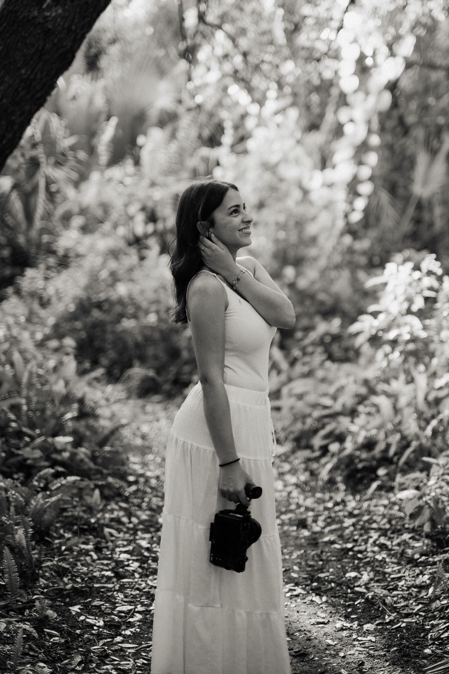 Woman in a white sleeveless top and long skirt smiles while holding a camera on a leafy forest path, in black and white.