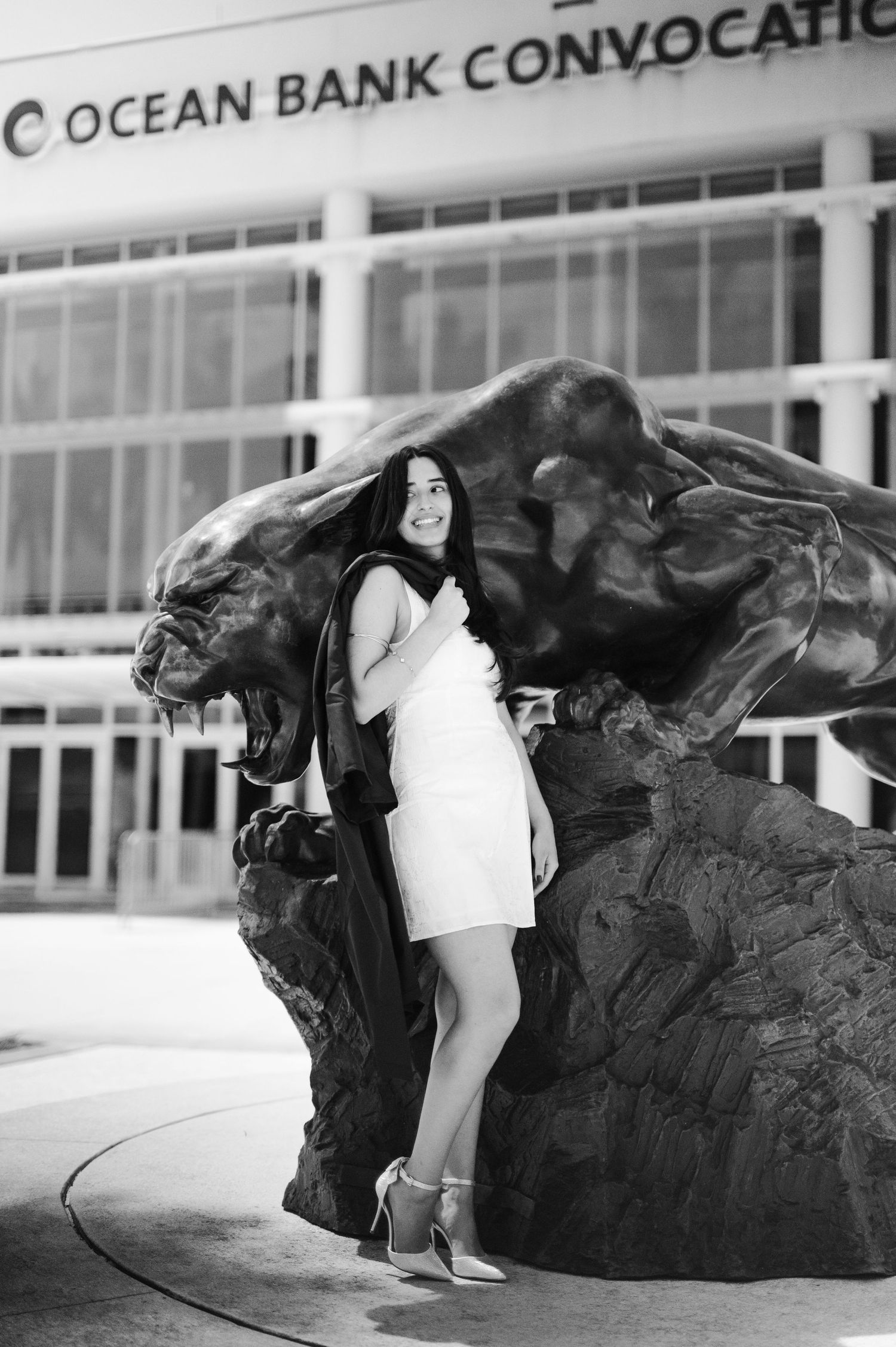 Black and white photo of a graduate in a white dress posing beside a large bronze panther statue outside a convocation center.