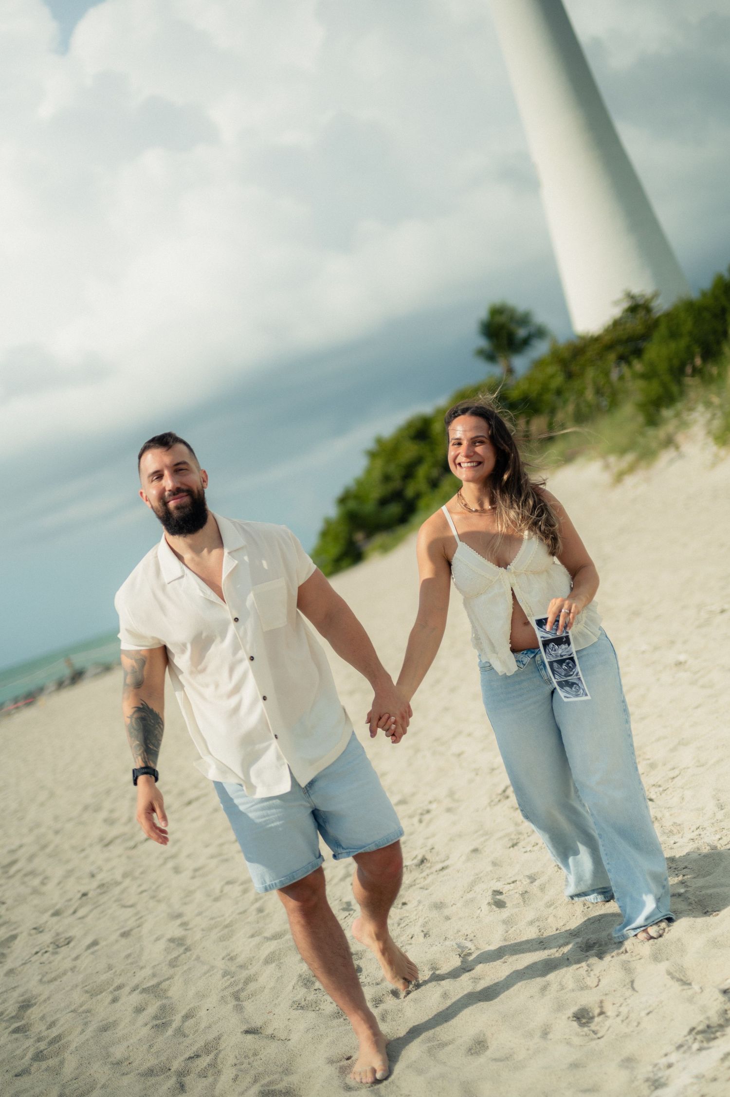 Happy couple holds hands and walks barefoot on a sandy beach with ultrasound photos, a lighthouse towering behind them.