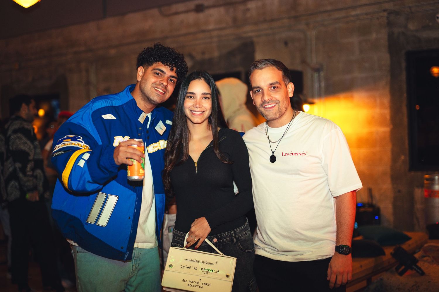Three friends smile for a photo at a lively evening gathering, one wearing a blue varsity jacket and holding a drink.