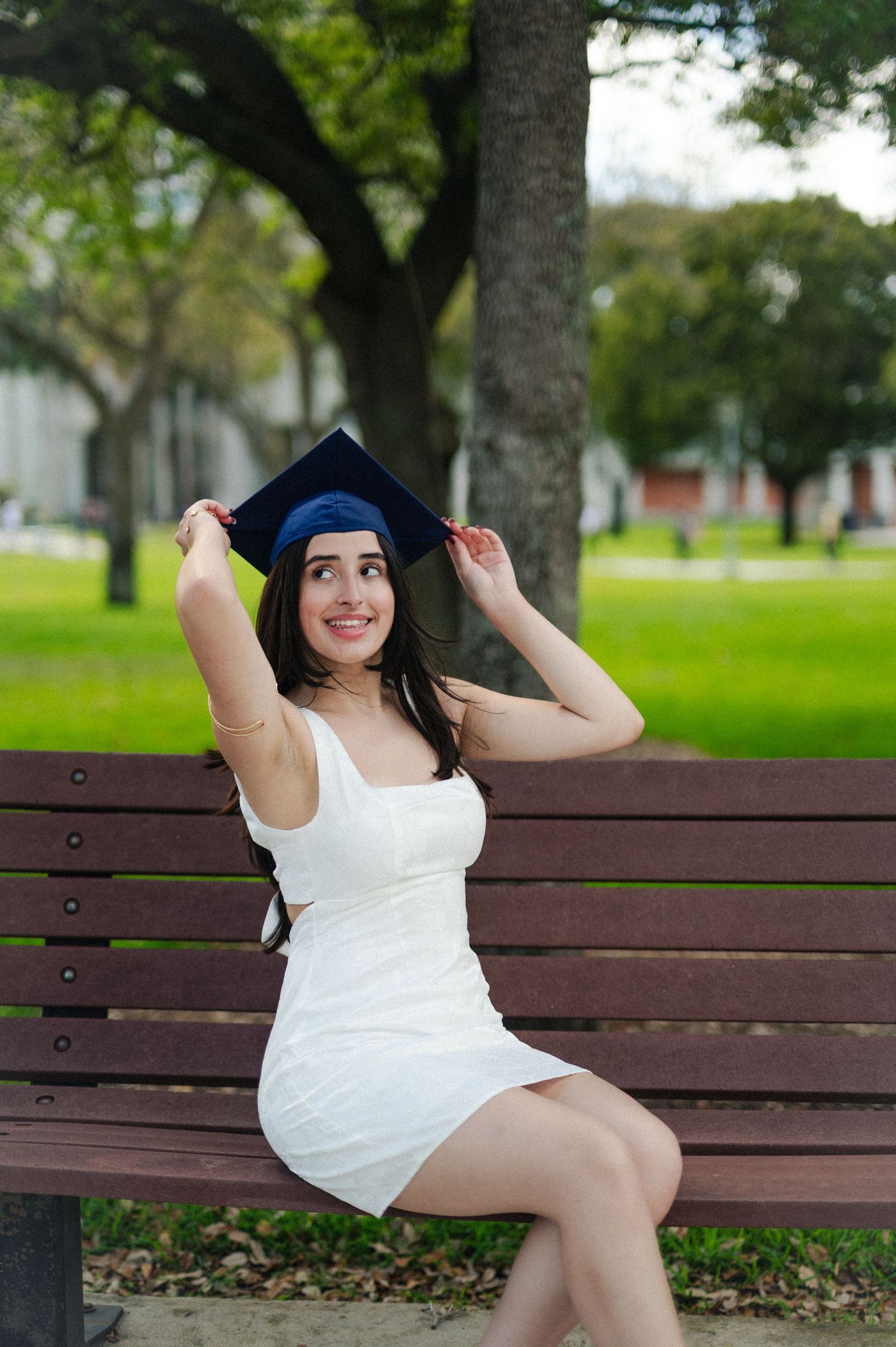 Woman in a white dress sits on a wooden park bench and adjusts a navy graduation cap on a sunny green campus.