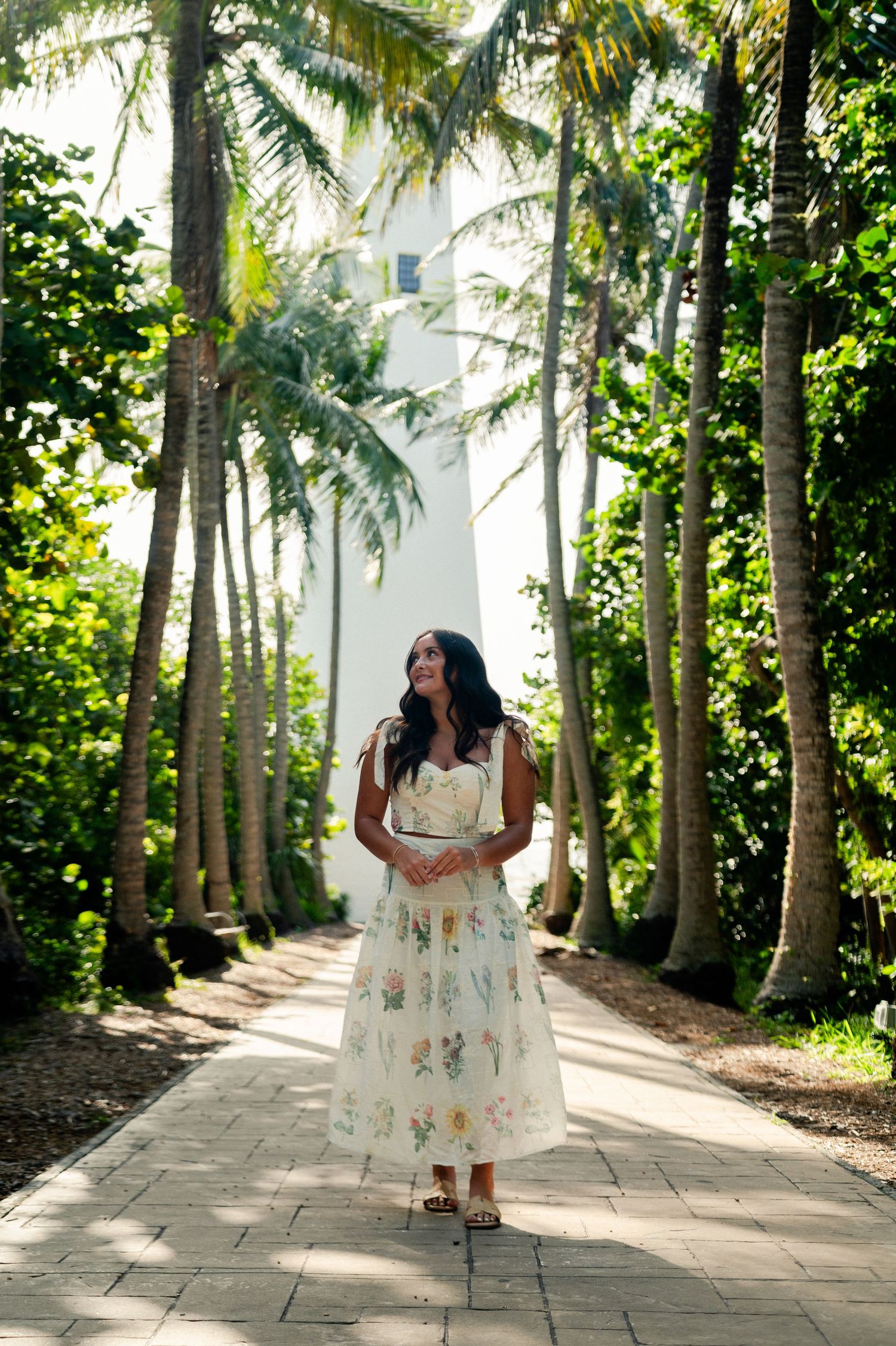 A woman in a floral cream sundress walks down a brick path lined with tall palm trees in a sunlit tropical garden.