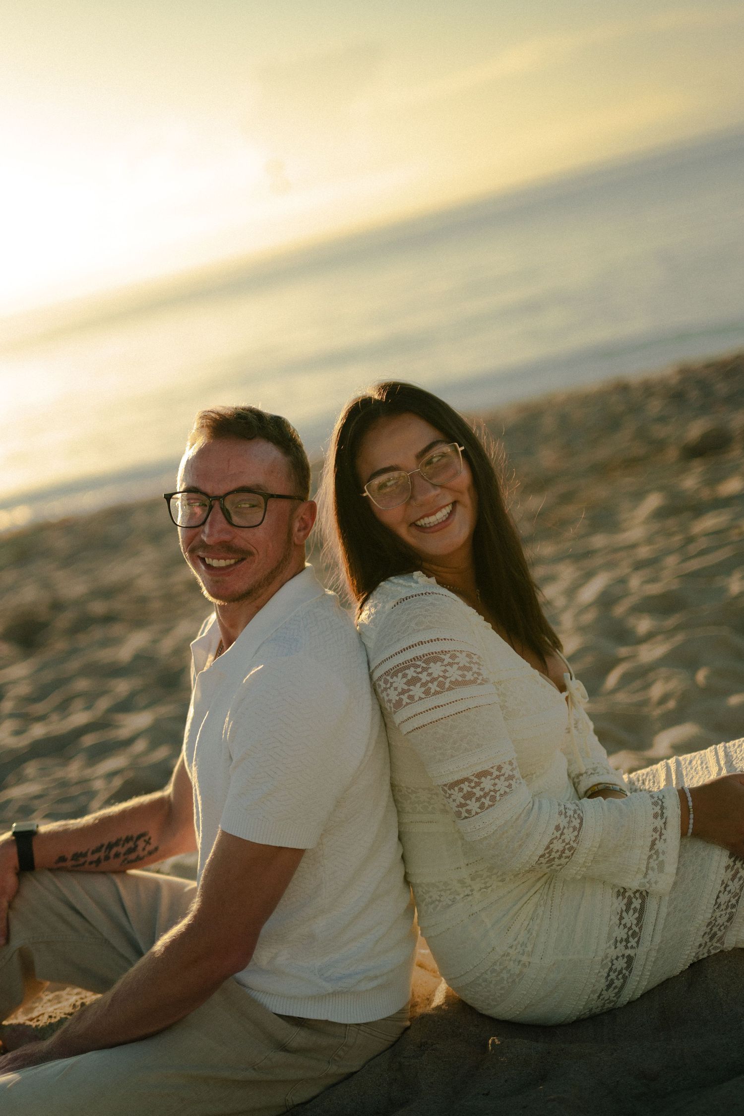 Smiling couple in glasses sit back to back on a beach at golden hour.