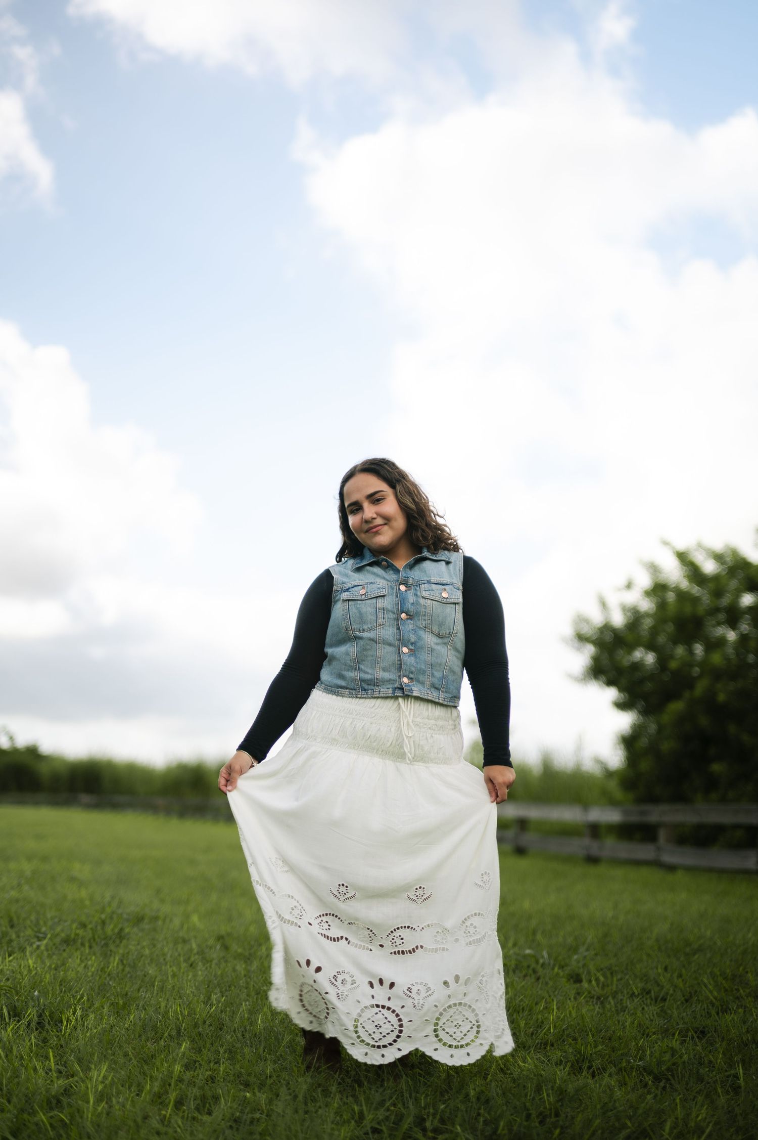 A smiling woman in a denim vest and white eyelet maxi skirt holds her skirt out while standing in a green field under cloudy skies.