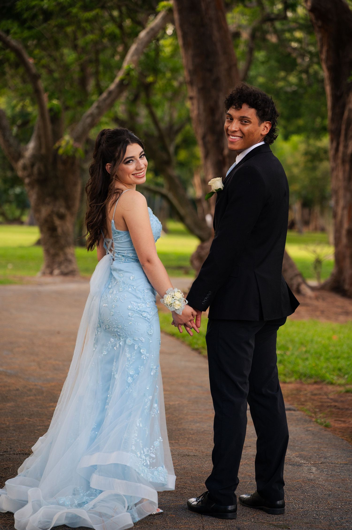A woman in a light blue lace gown and a man in a black suit hold hands and smile back at the camera on a tree-lined park path.