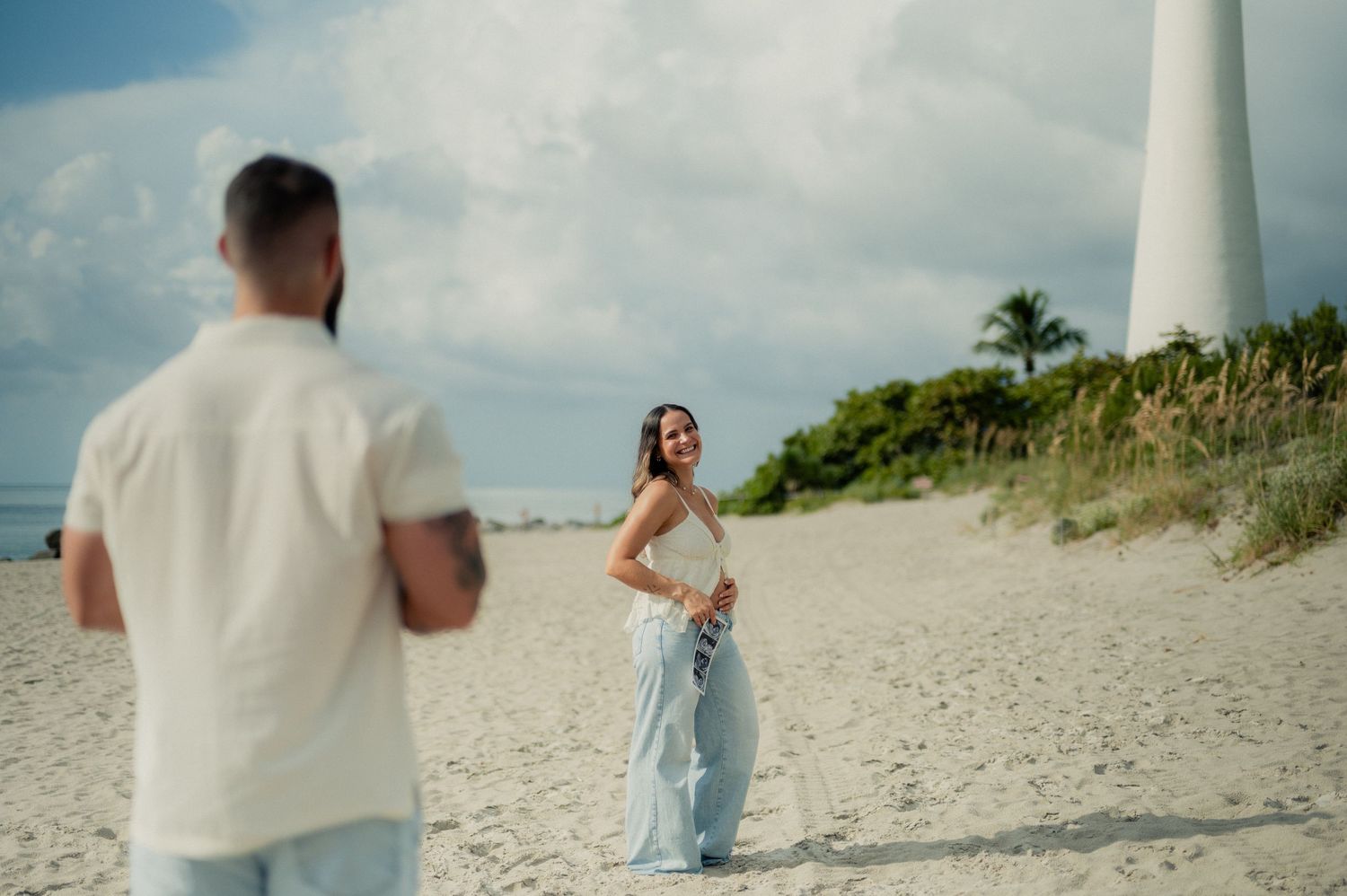 Woman holding ultrasound photos smiles on the beach while her partner stands in the foreground near a white lighthouse.
