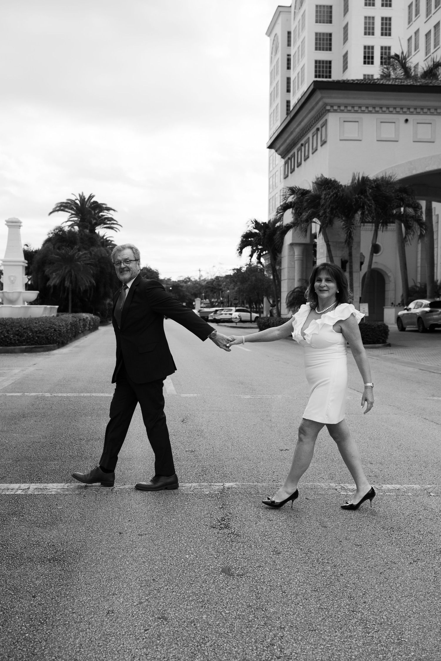 Black and white photo of a couple holding hands and walking across a palm-lined street in front of a building.